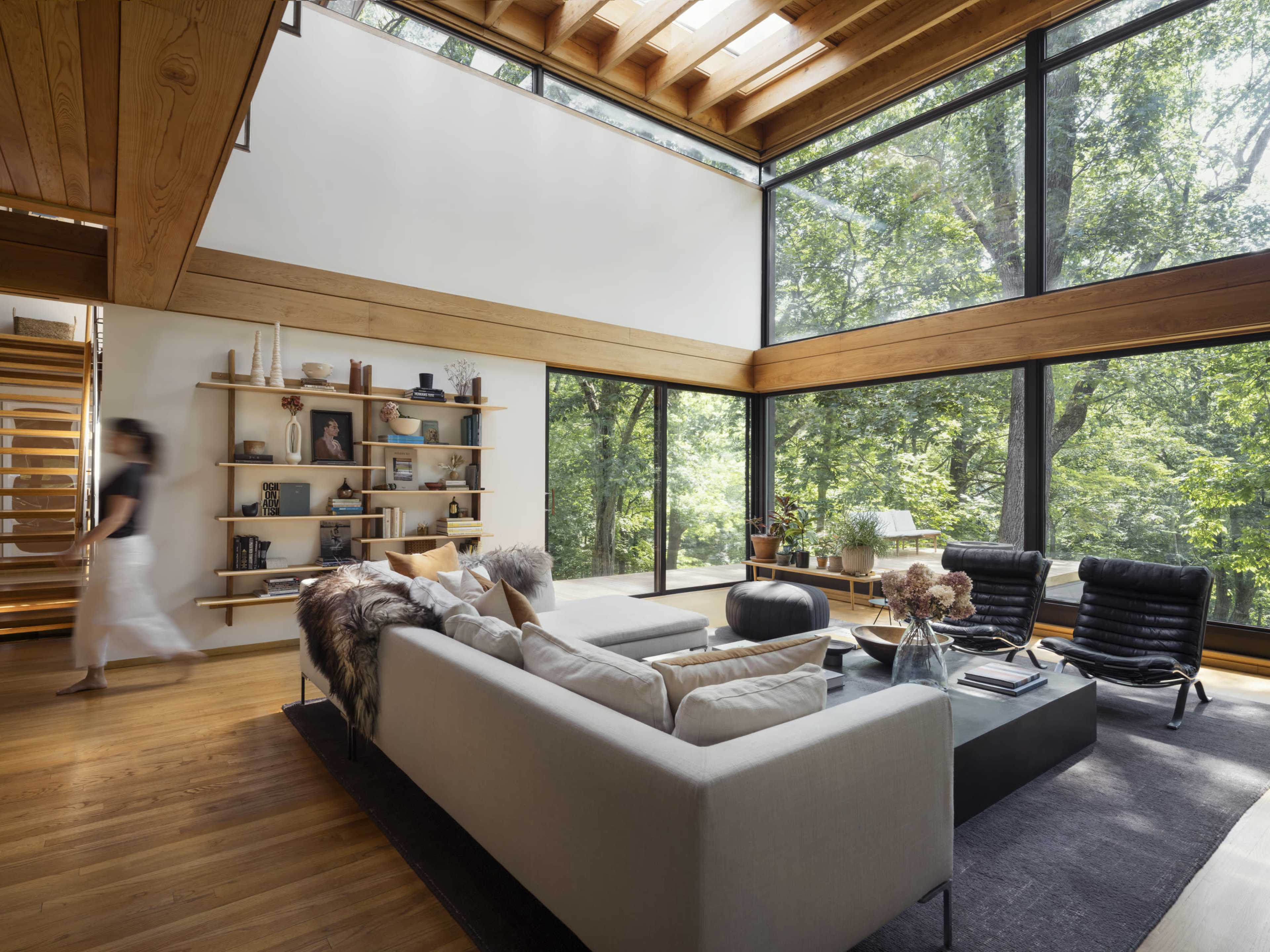 A modern living room features large windows with a view of trees, a sectional sofa, and minimalist shelving.