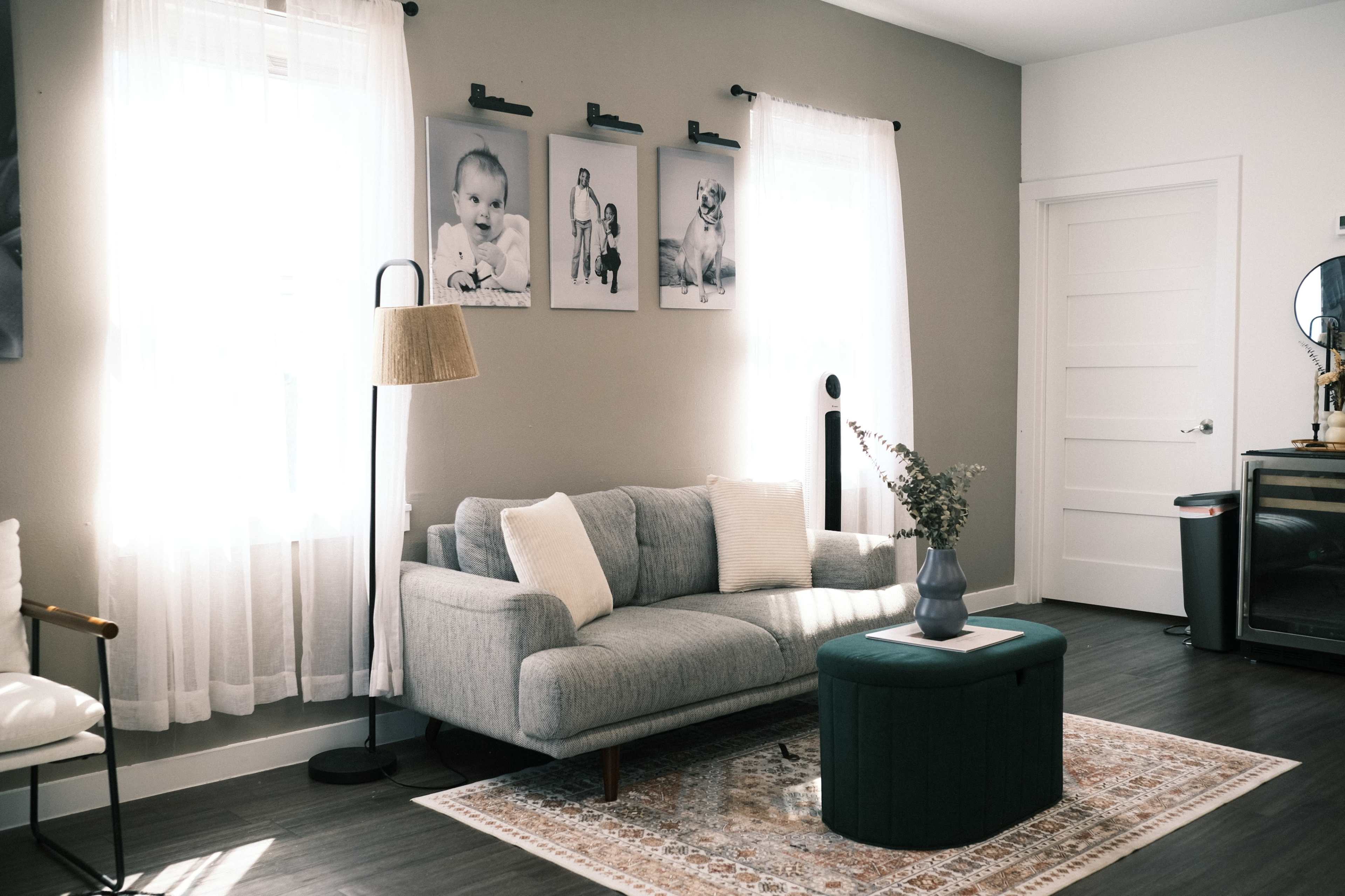 The room features a light-filled space with a gray sofa, a green ottoman, and three black-and-white framed photographs on the wall, complemented by a lamp and a decorative vase.