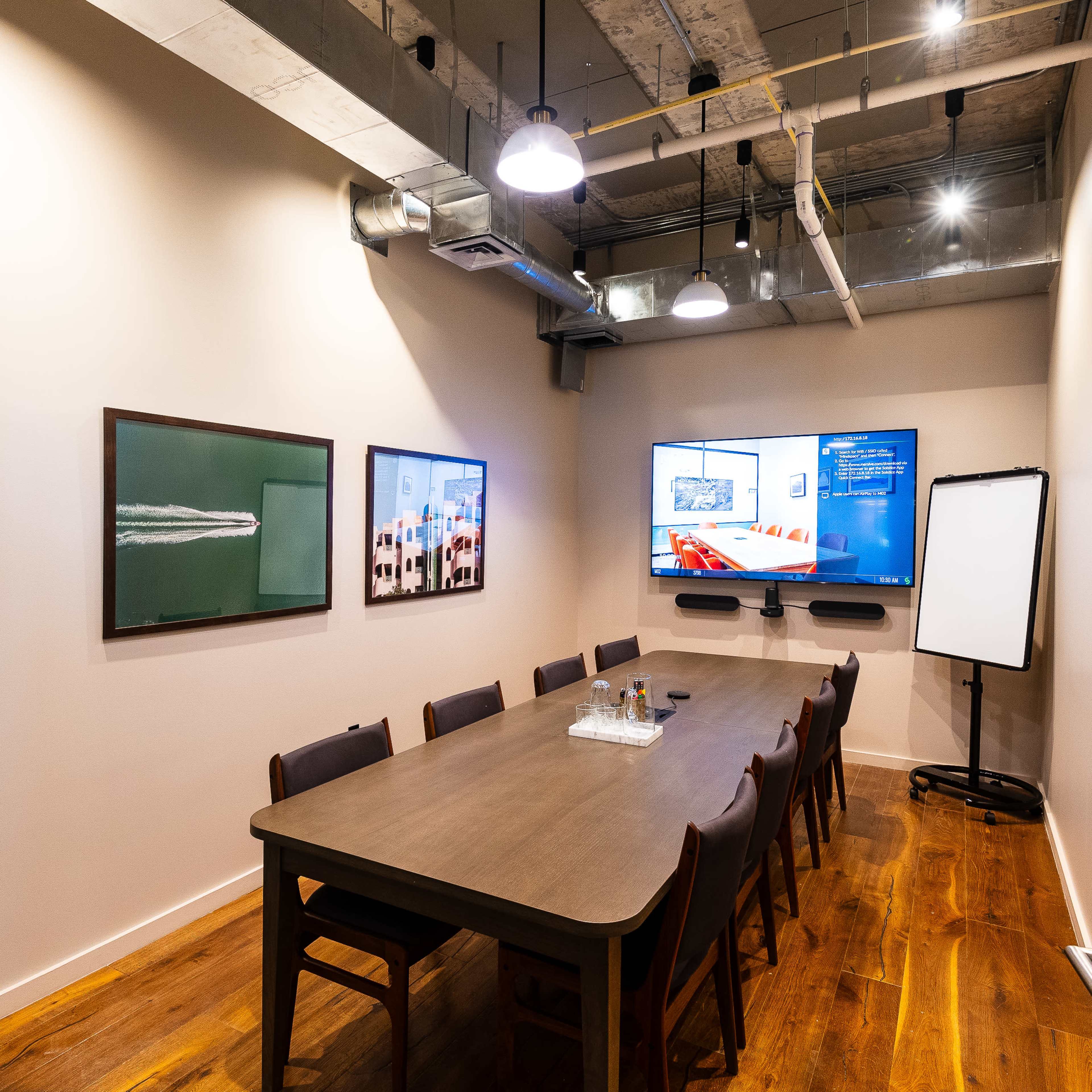 A well-lit conference room features a long wooden table surrounded by chairs, with two framed artworks on the walls and a large screen displaying information.