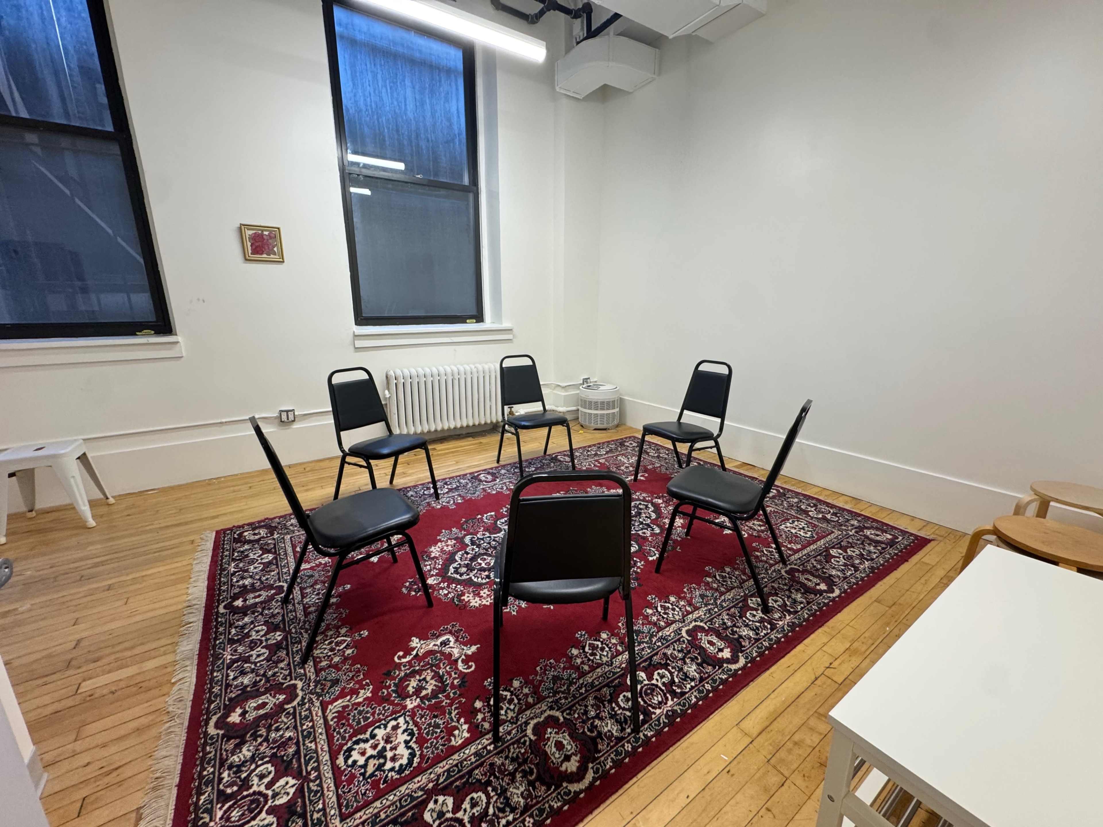 The image shows a small, sparsely furnished room with six black chairs arranged in a circle on a red rug.
