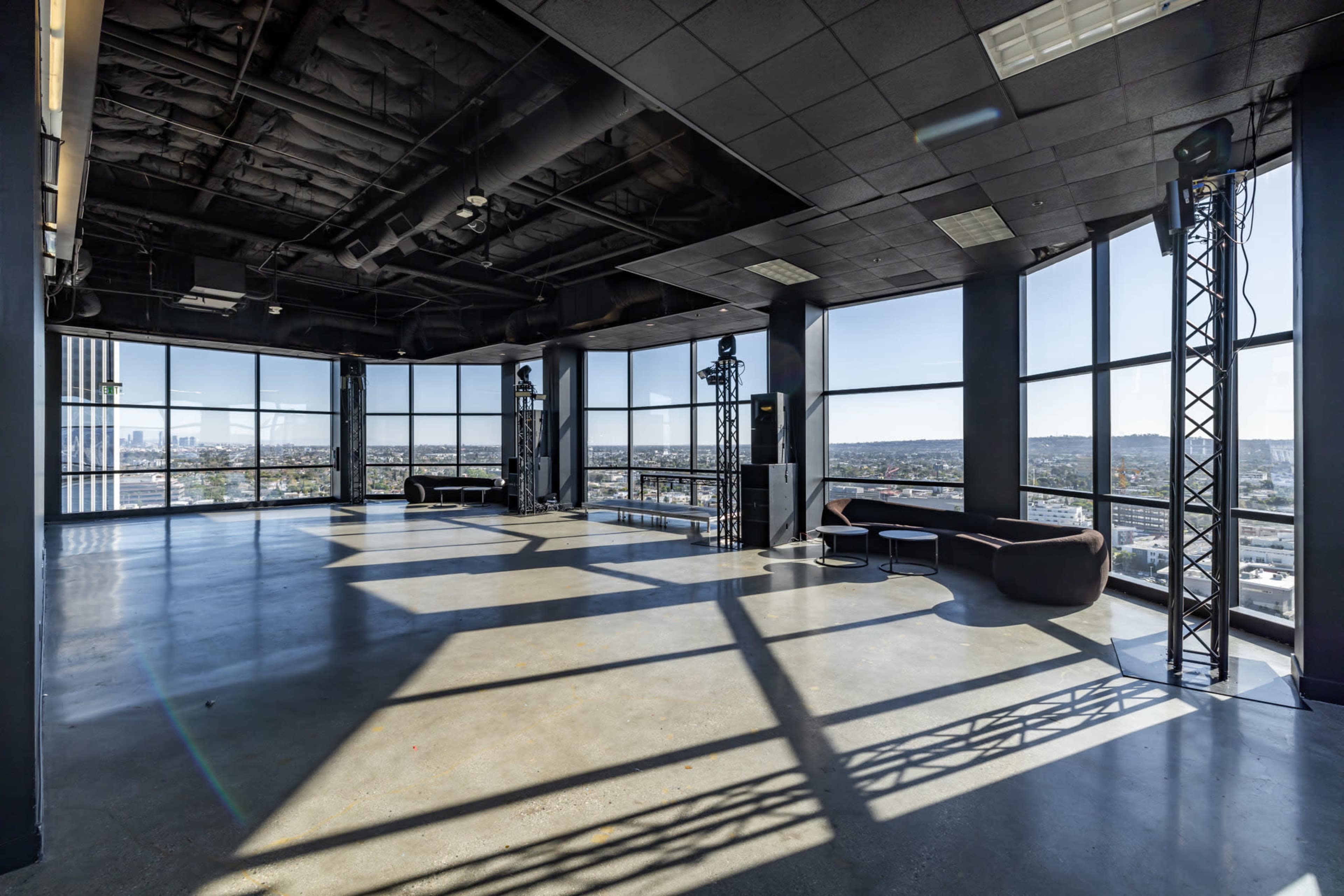 The image shows a spacious, modern interior with large windows, concrete floors, and minimal furniture, offering a panoramic view of the city.