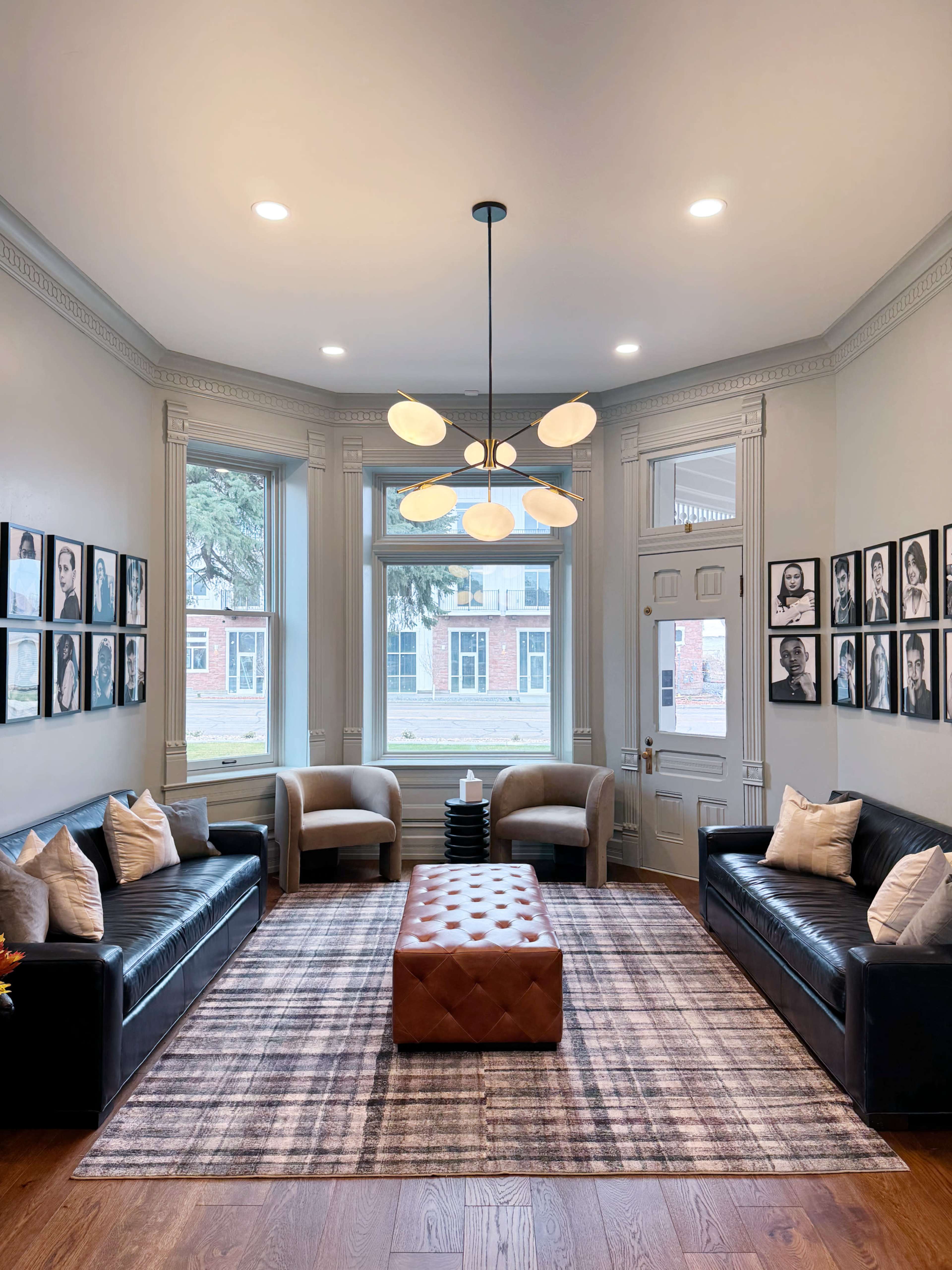 A modern living room with two leather sofas, a large brown ottoman in the center, and a series of framed portraits on the walls, all surrounded by large windows.