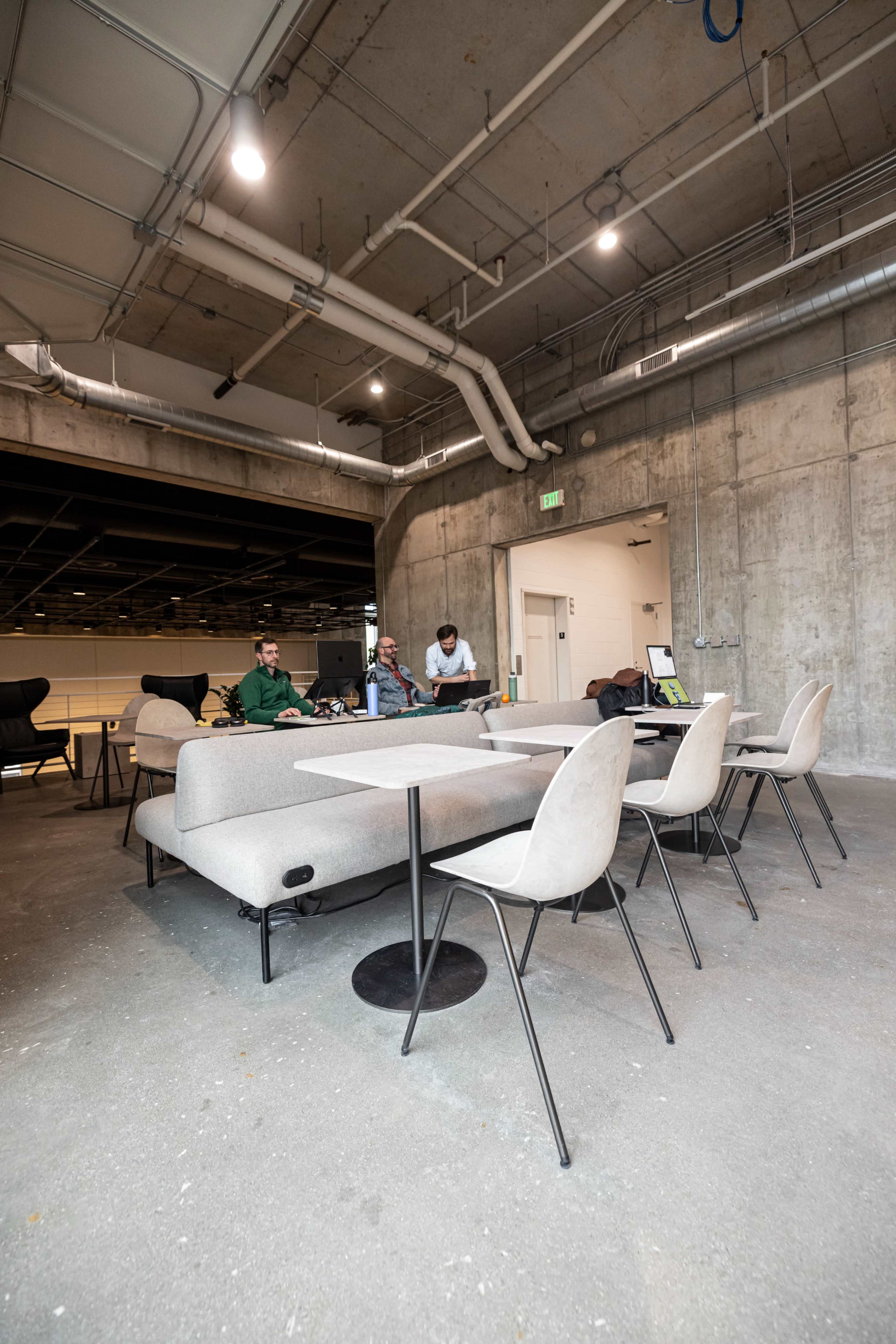 The image depicts a modern, industrial-style workspace featuring three people gathered around a table with light gray sofas and minimalist chairs in a concrete-finished area.