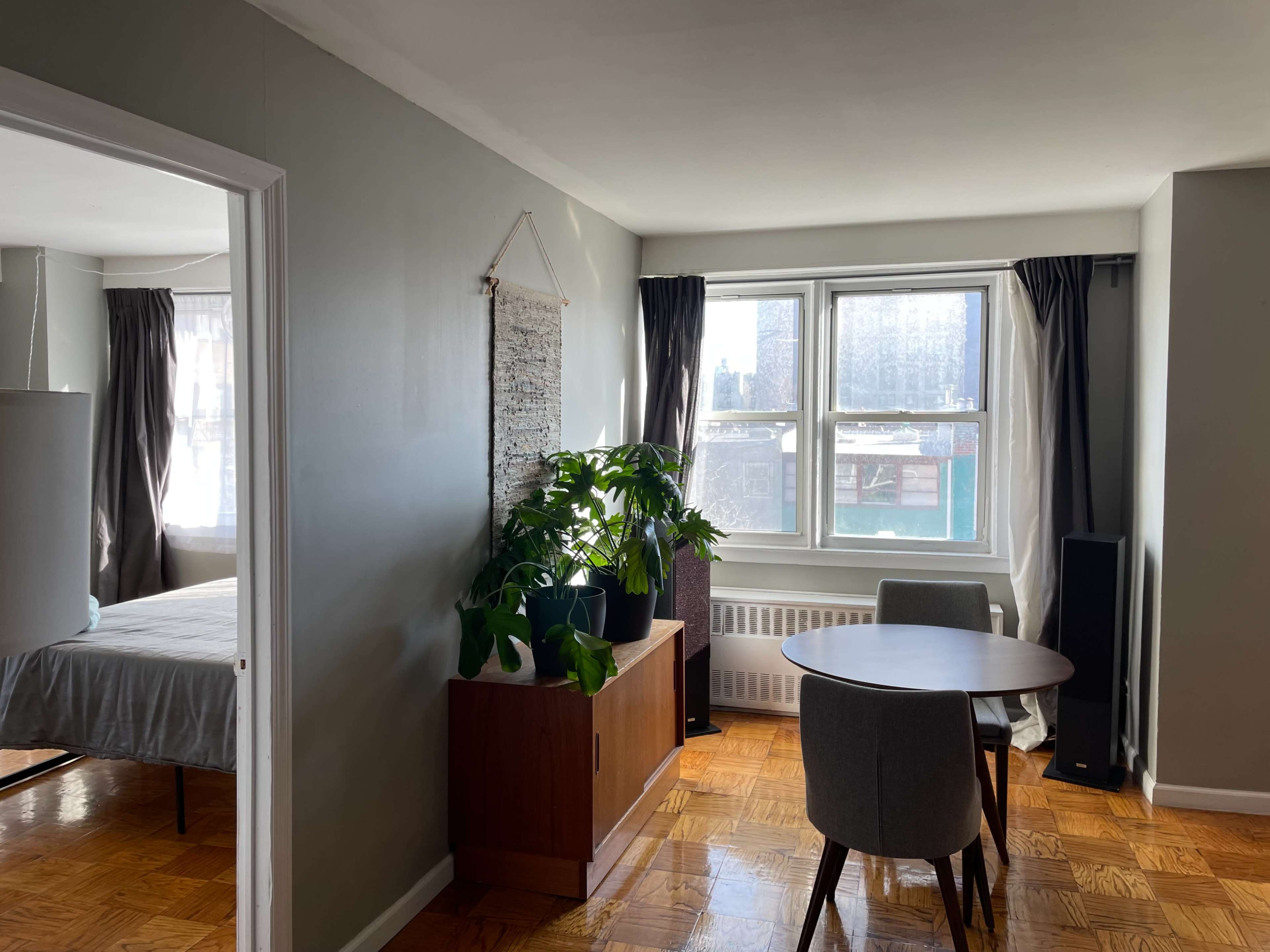 The image shows a well-lit room with a dining table and chairs, a potted plant, and a view of a bed in a separate area.