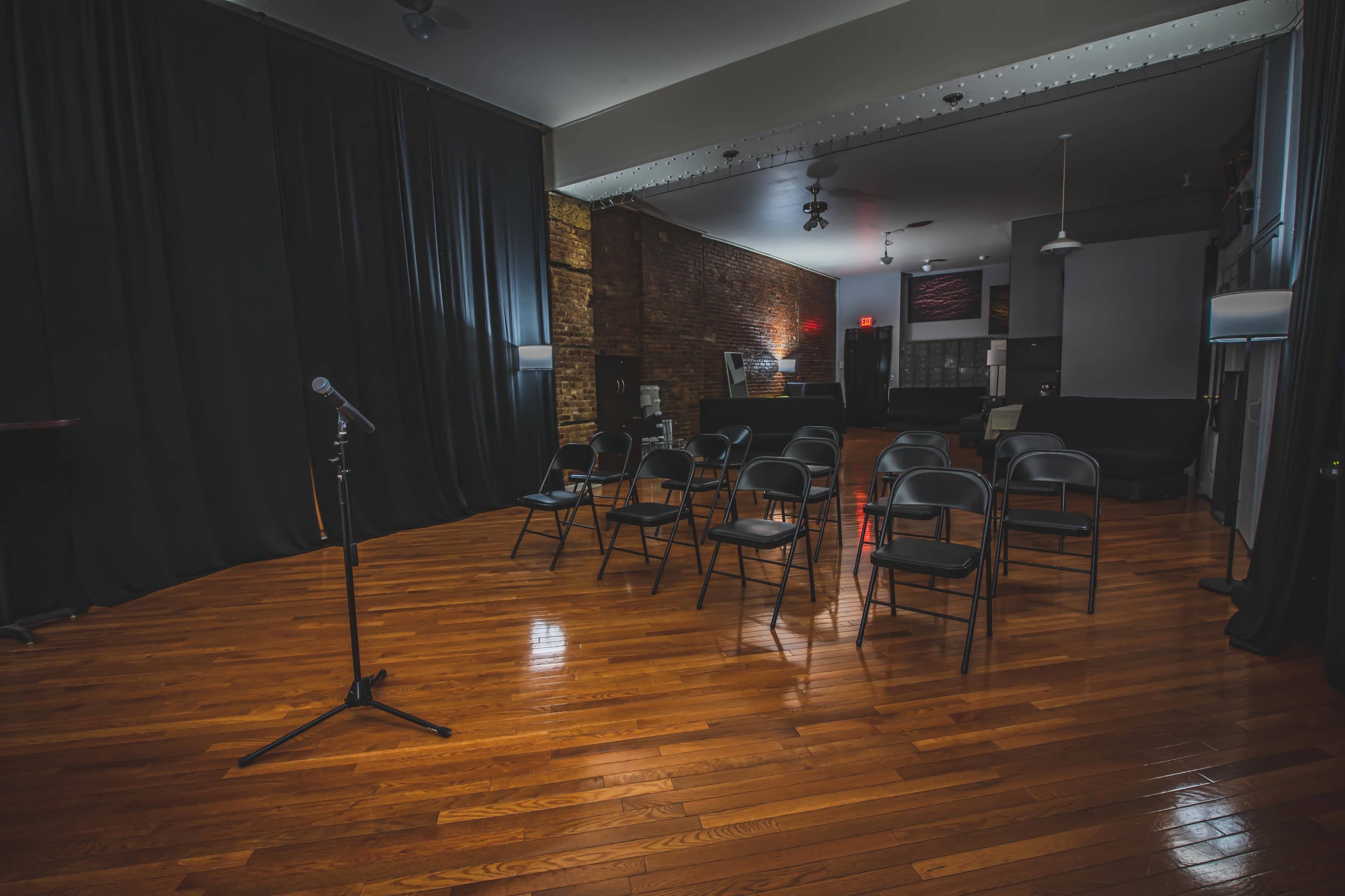 A small performance space features a microphone on a stand in front of several empty black chairs arranged in a semicircle on a polished wooden floor.