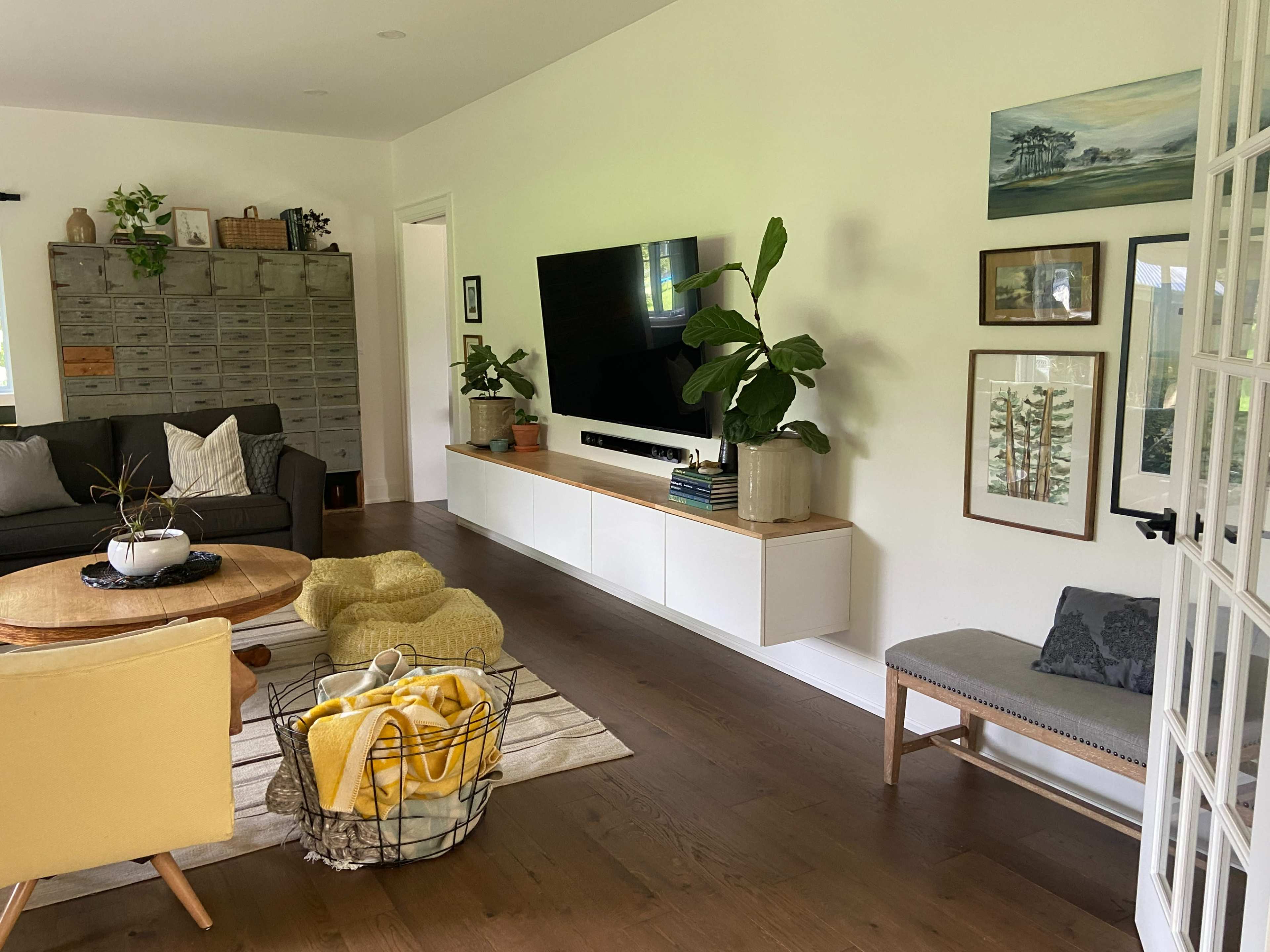 The image shows a modern living room with a sectional sofa, a coffee table, a wall-mounted television, and decorative plants, alongside a side table and a shelving unit.