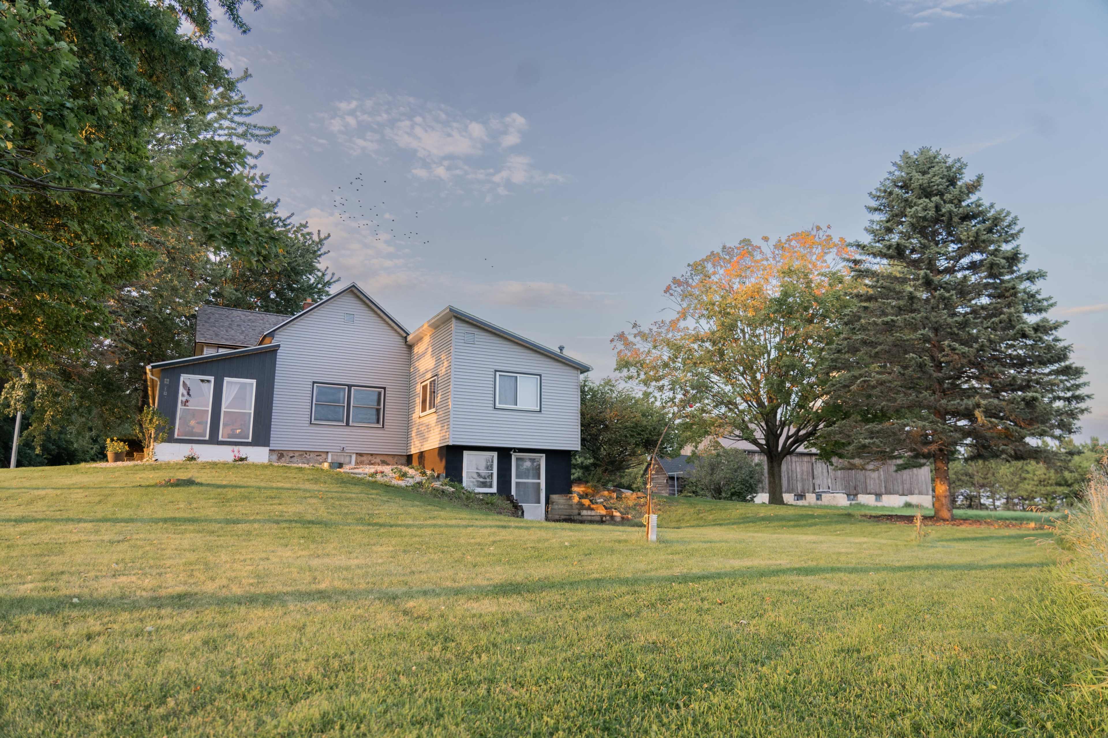 A gray two-story house sits on a grassy hill surrounded by trees and clear skies.