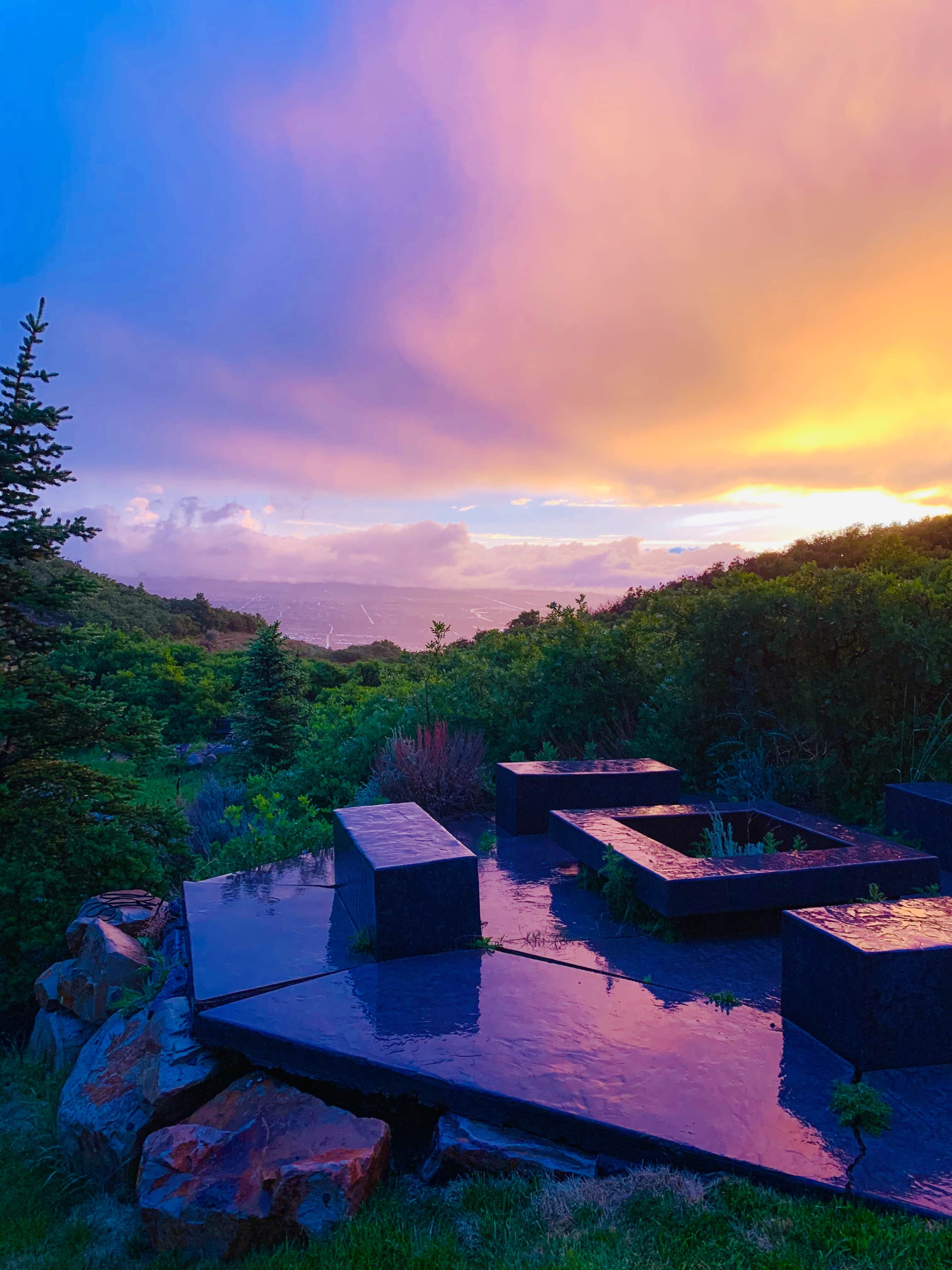 A dark stone patio with rectangular seating overlooks a landscape of trees and hills under a colorful sunset sky.