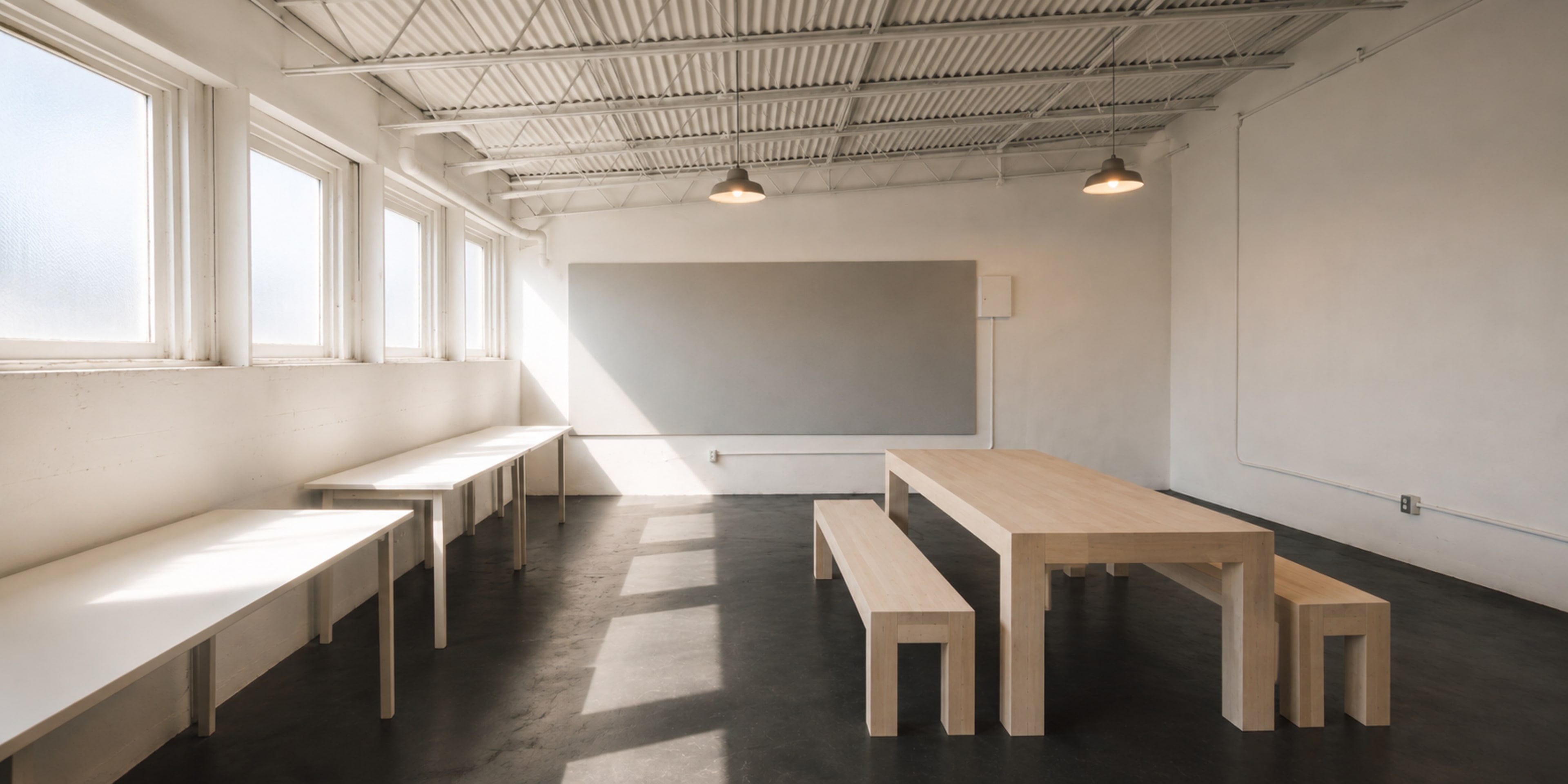 The image shows a minimalist room with large windows, a long wooden table in the center, and benches on either side, along with two narrow side tables against the walls.