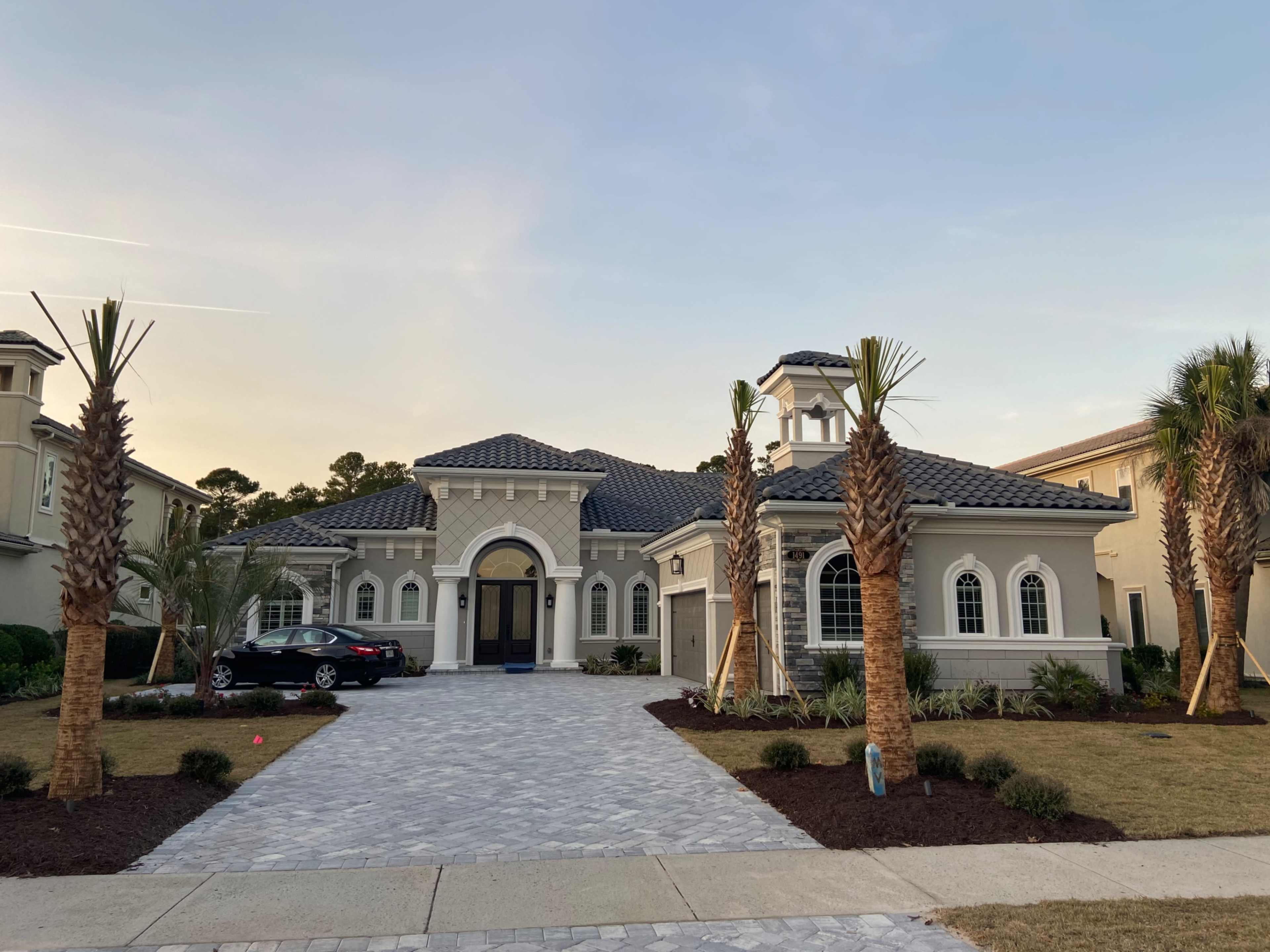 A large, modern home with a curved driveway is flanked by palm trees and features a stone pathway leading to the entrance.