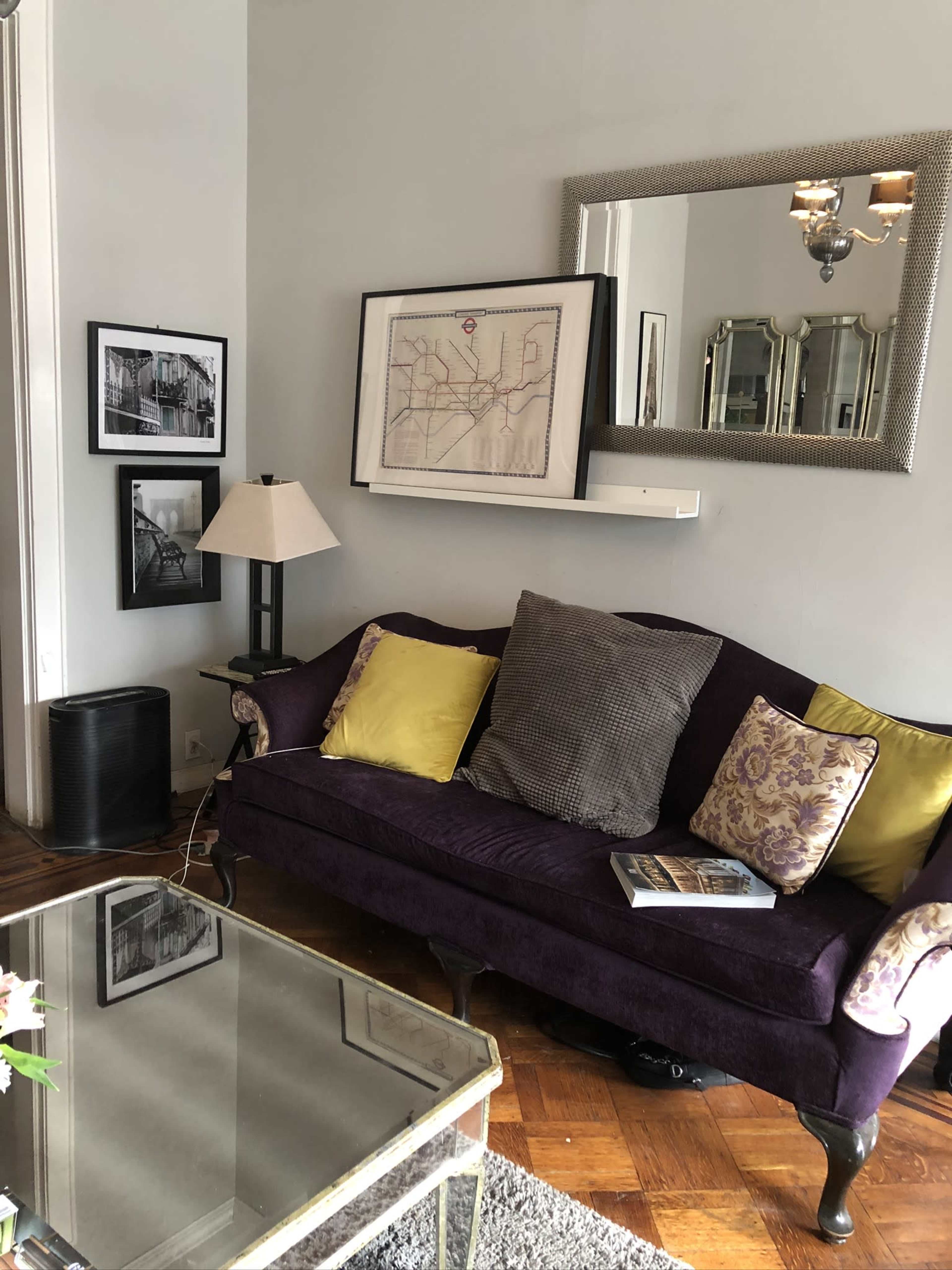 A purple sofa with decorative pillows sits next to a coffee table in a well-decorated living room featuring framed photographs and a wall mirror.