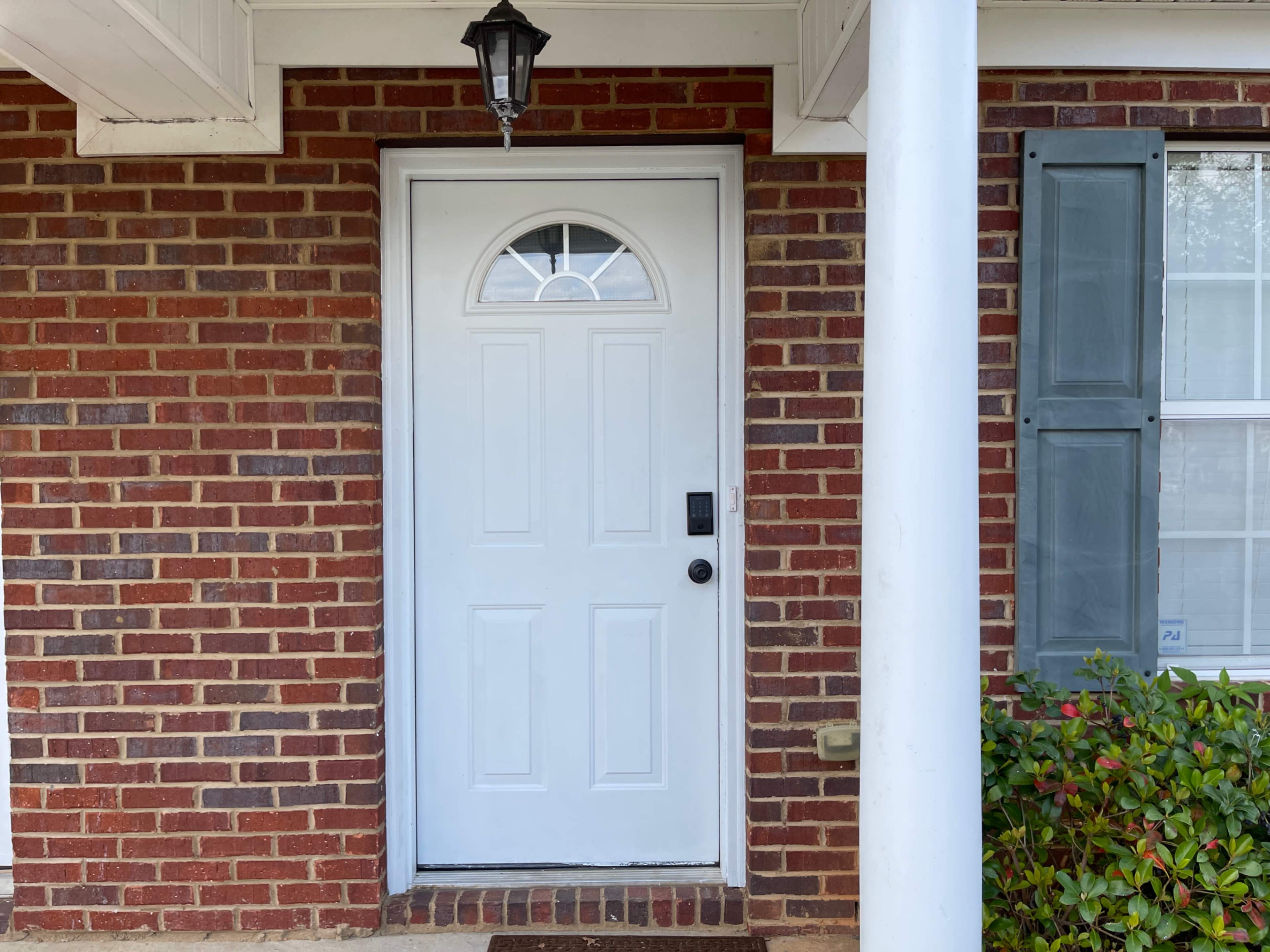 The image shows a white front door with an arched window, set against a brick wall with green shutters on either side.