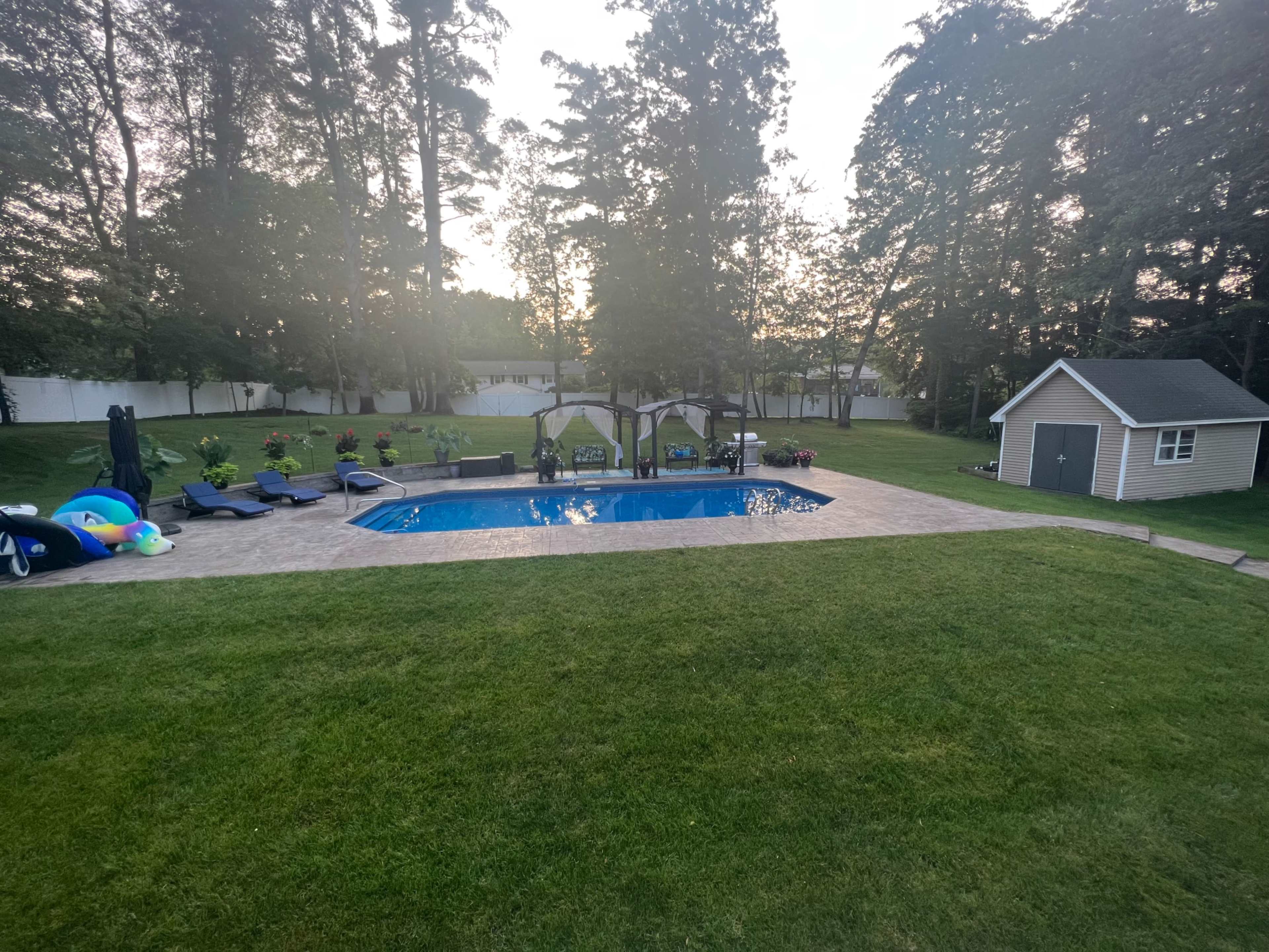 The image shows a backyard with a swimming pool surrounded by lounge chairs, shaded seating areas, and a small shed.