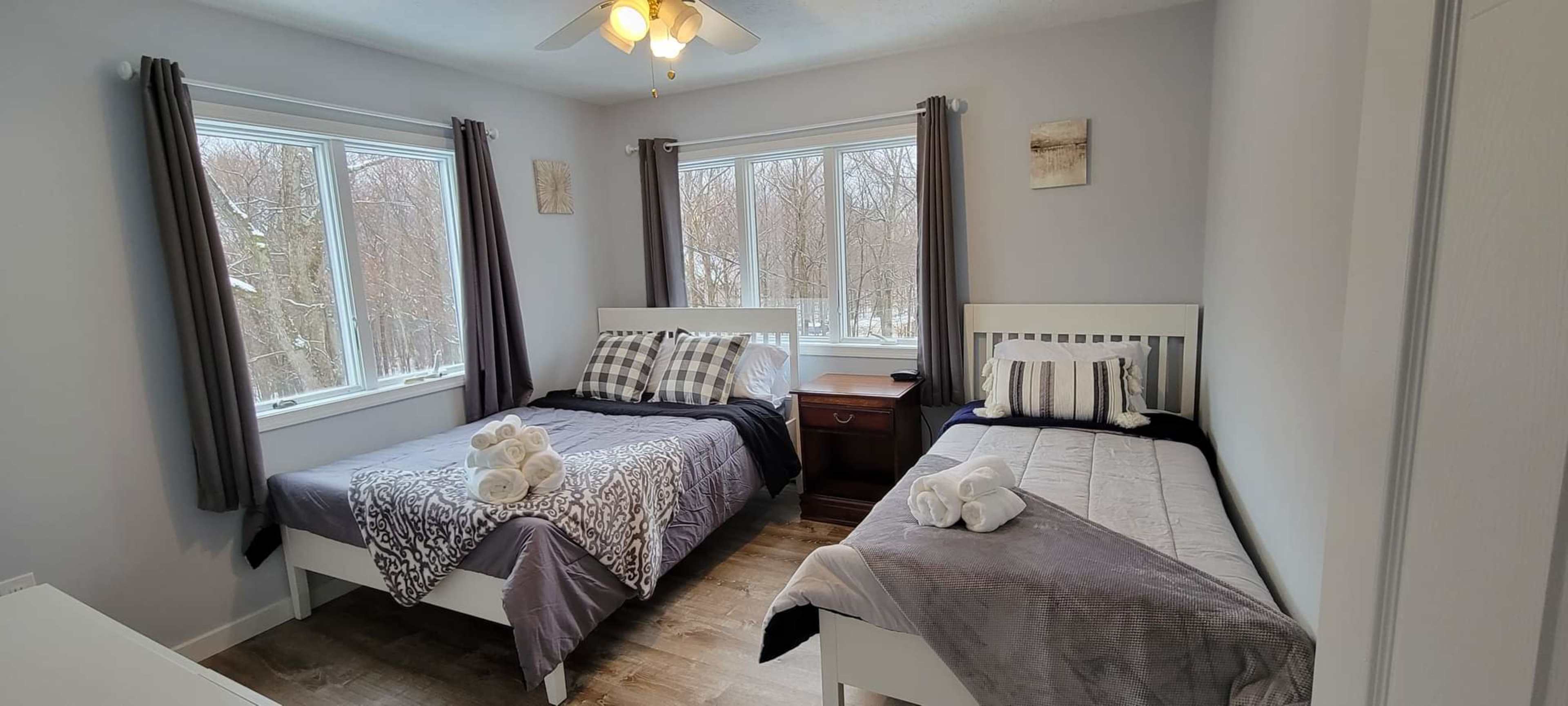 The image shows a well-lit bedroom with two beds, each neatly made with gray and white bedding, and a nightstand between them.