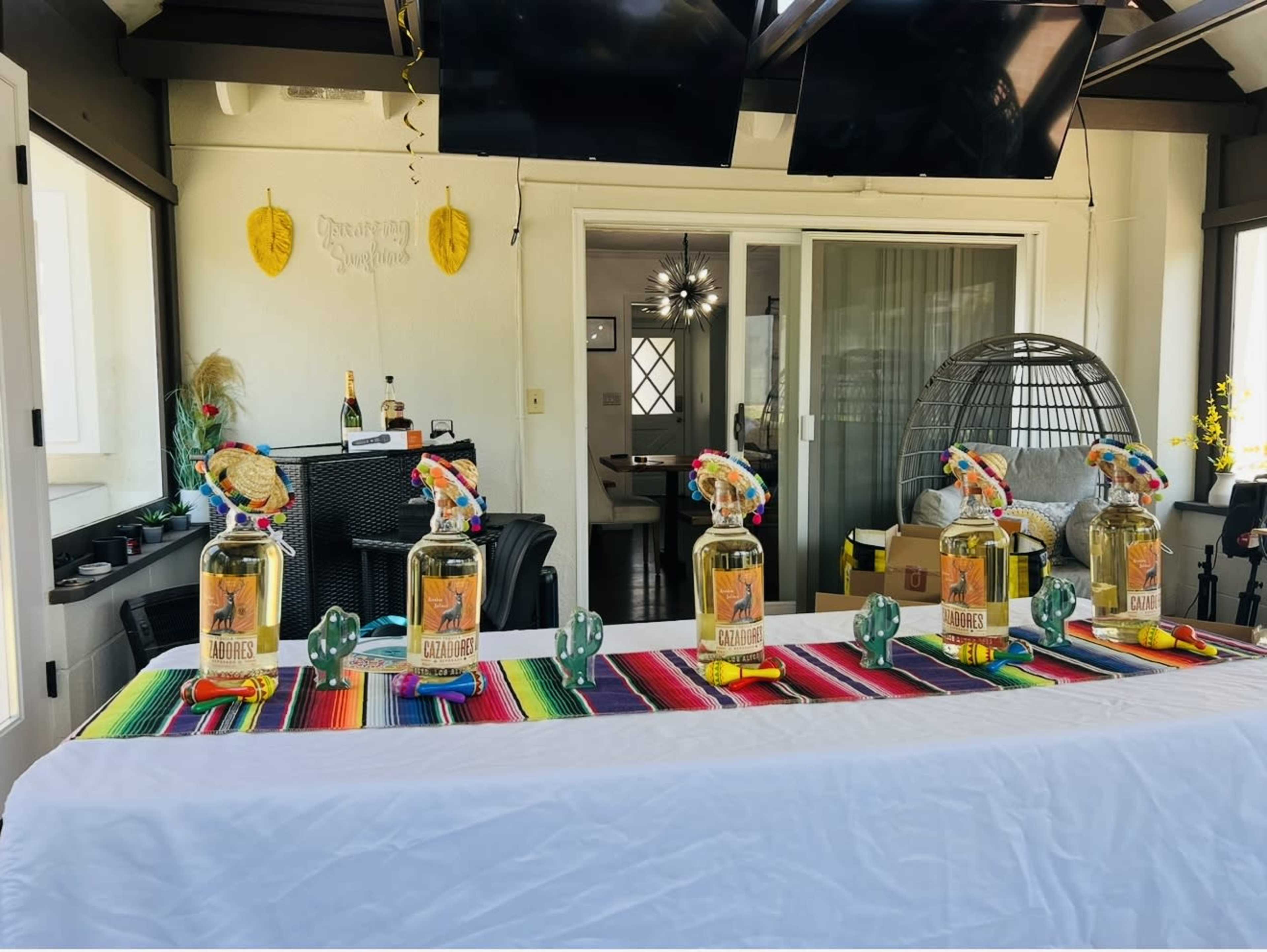 A colorful table is set up with five bottles of tequila arranged on a vibrant striped blanket, surrounded by decorative items, in a well-lit room with windows and modern furniture.