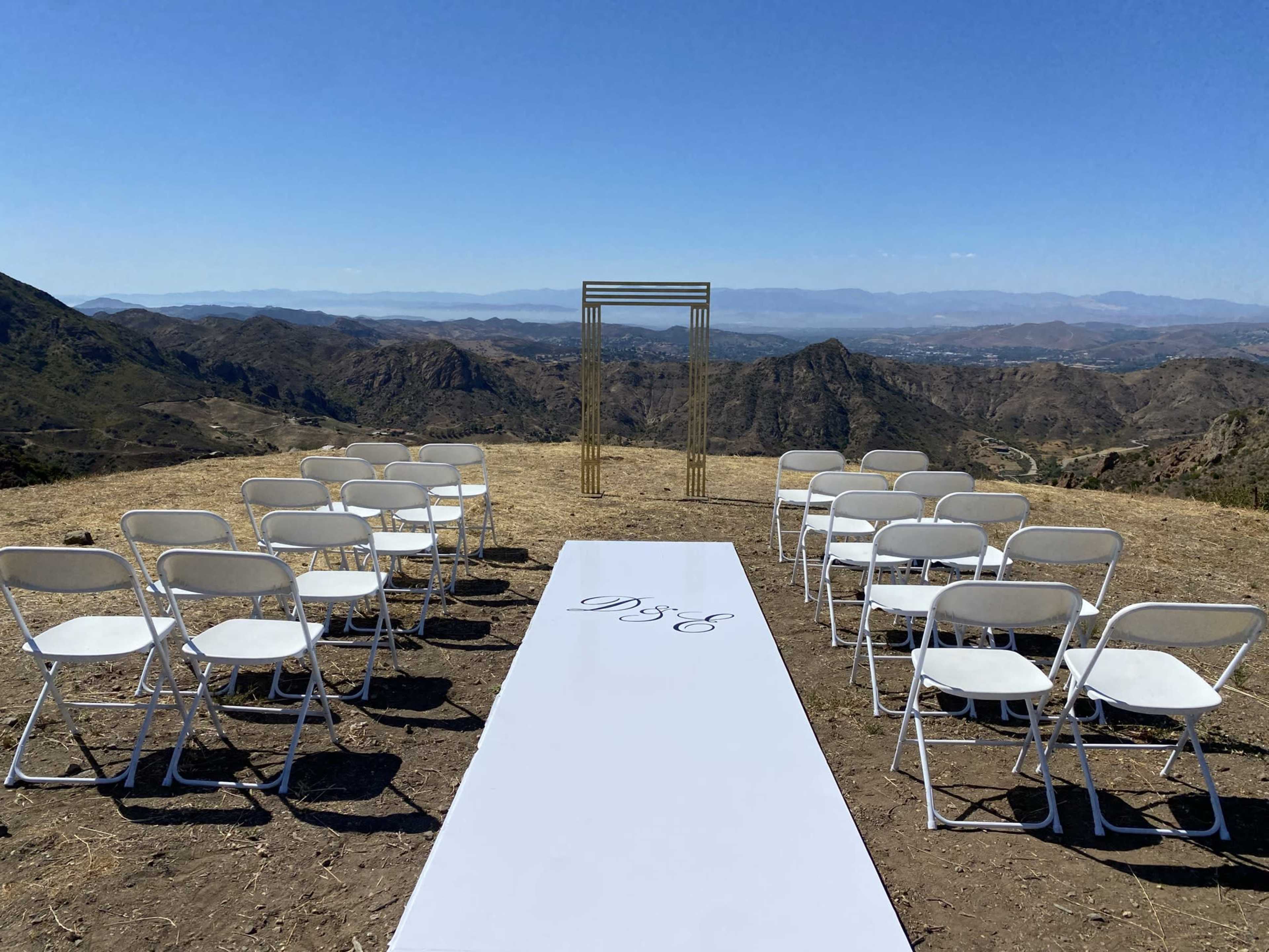 An outdoor wedding ceremony setup features white chairs arranged on a grassy area with a simple arch in the background, overlooking a mountainous landscape.