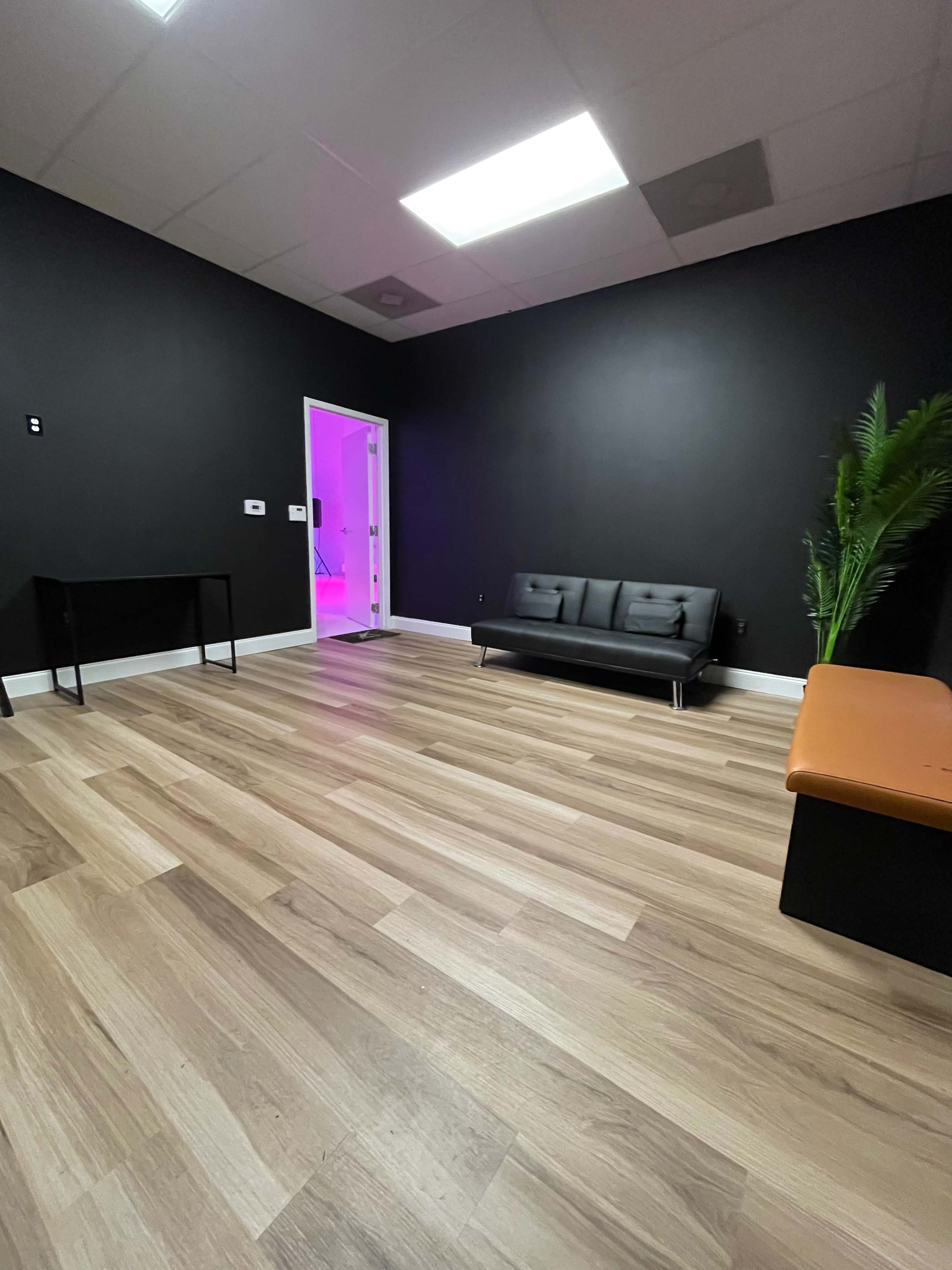 A sparsely furnished room with a dark wall, a black sofa, and light-colored wooden flooring.