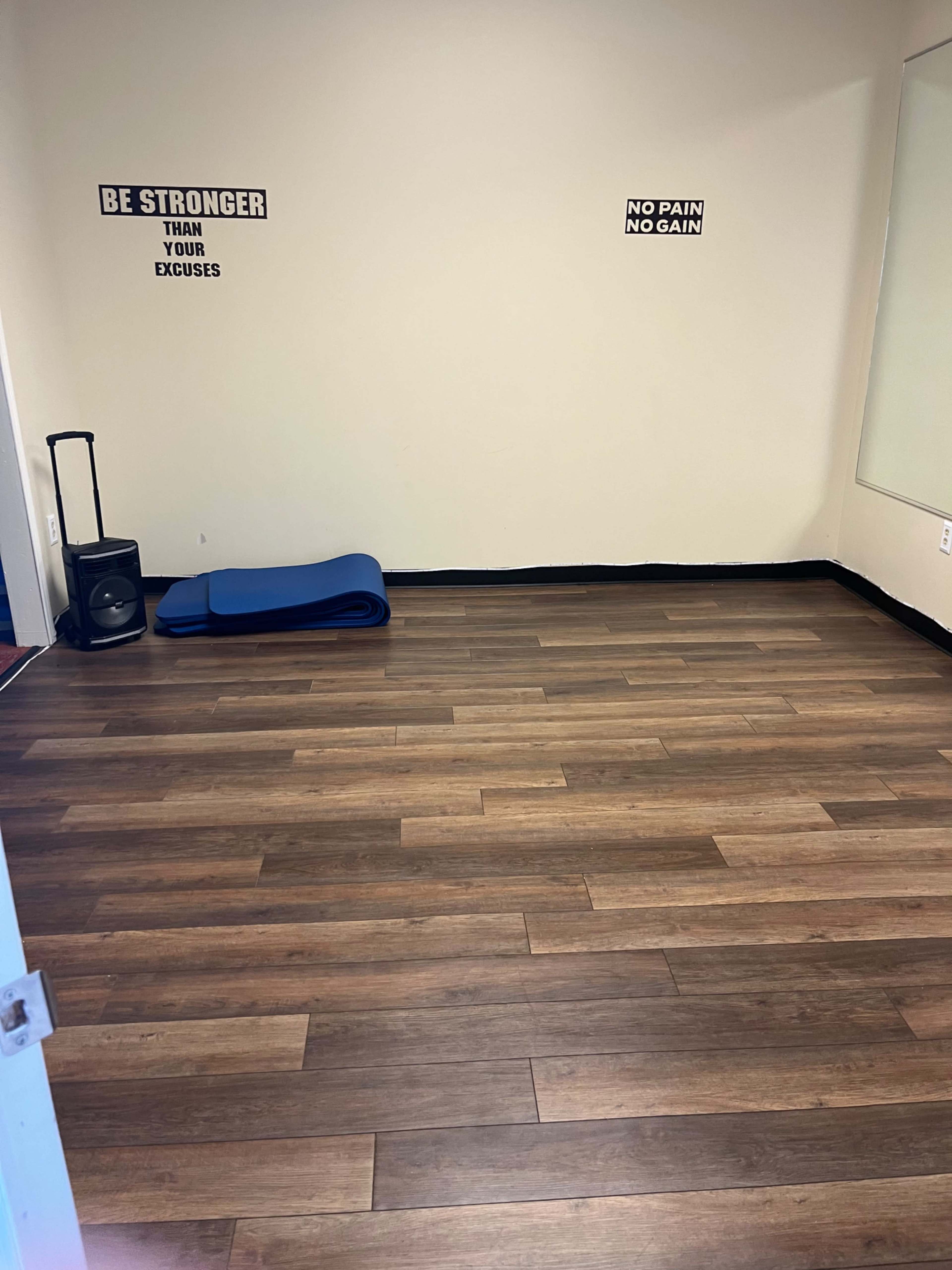 The image shows a sparsely furnished exercise room with wooden flooring, a blue mat on the ground, and motivational signs on the walls.