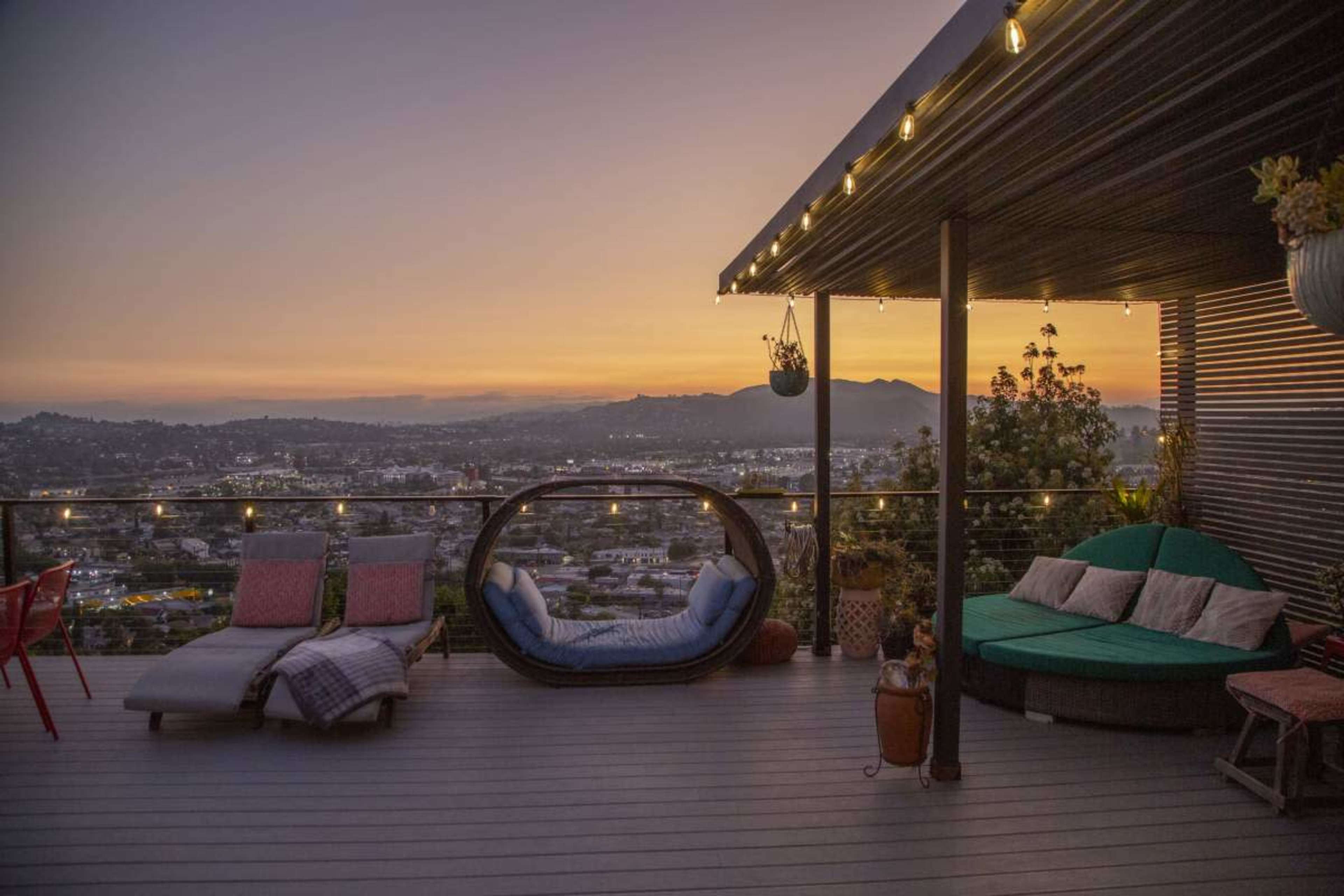 The image shows a modern rooftop patio with a cozy seating area and a view of a city skyline at sunset.