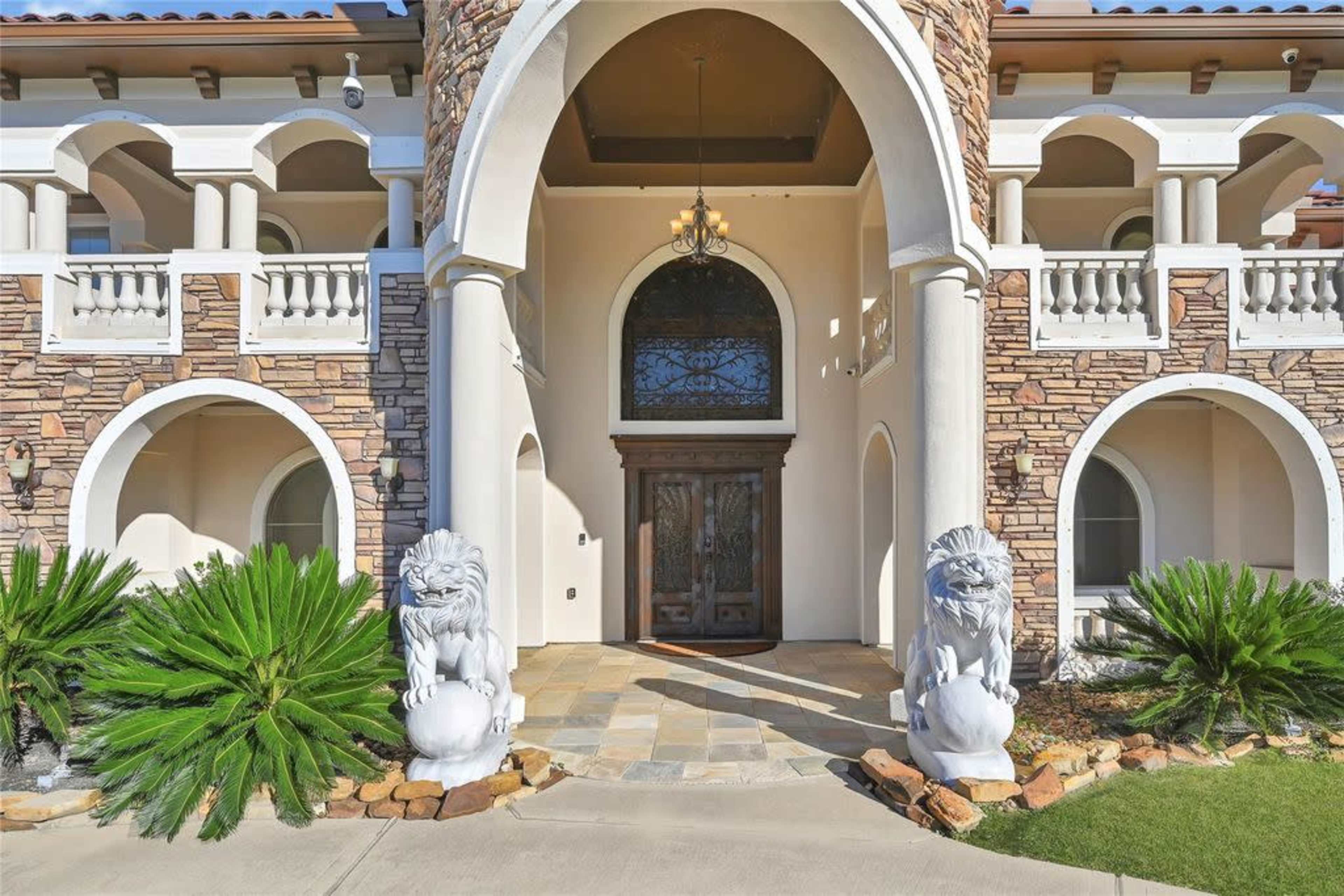 The image shows a large, ornate entrance of a house featuring a stone facade, two lion statues flanking the doorway, and a decorative arch above the entrance.