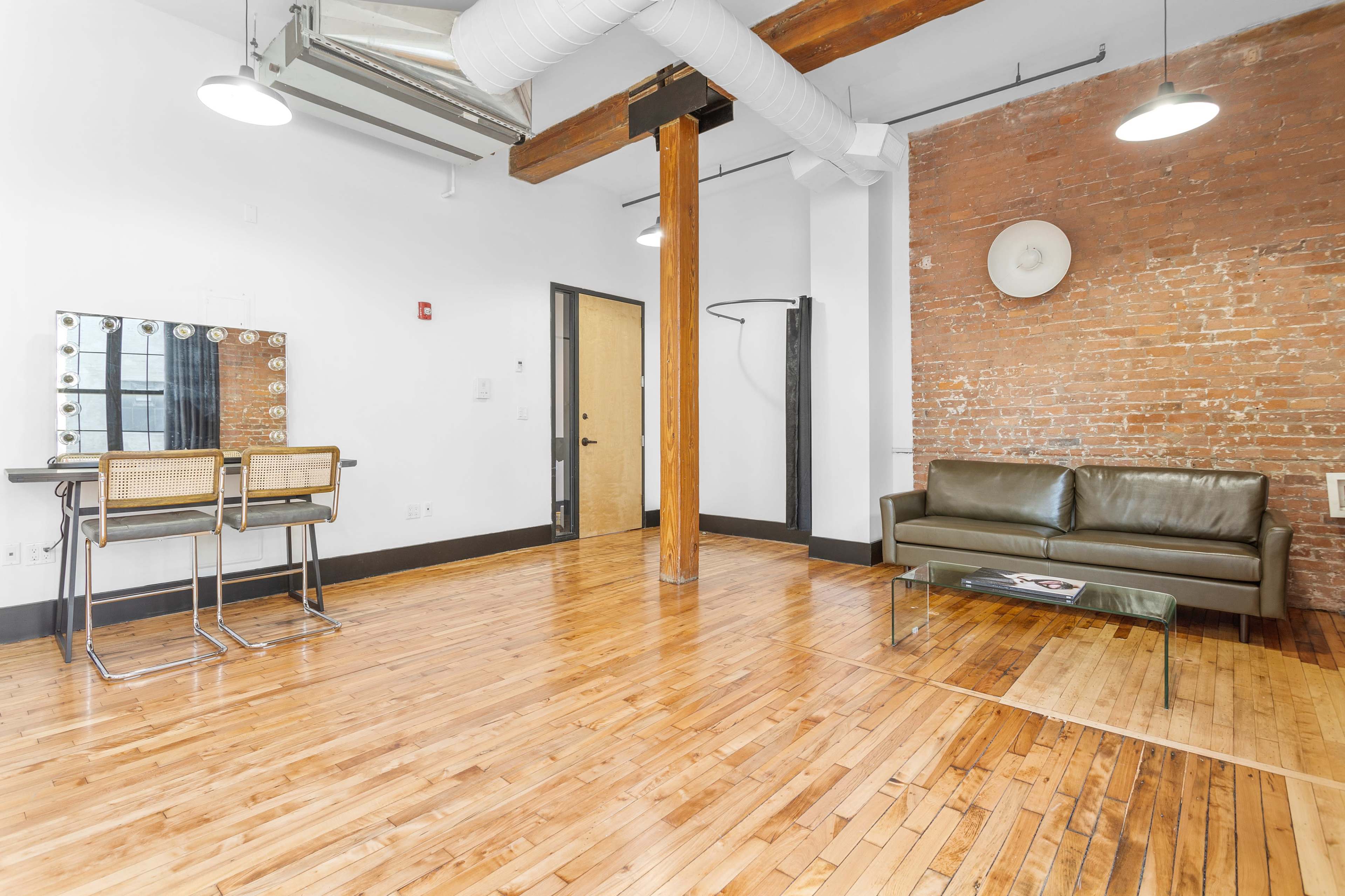 A modern interior with exposed brick walls, wooden floors, a green sofa, a glass coffee table, and a mirrored vanity with two stools.
