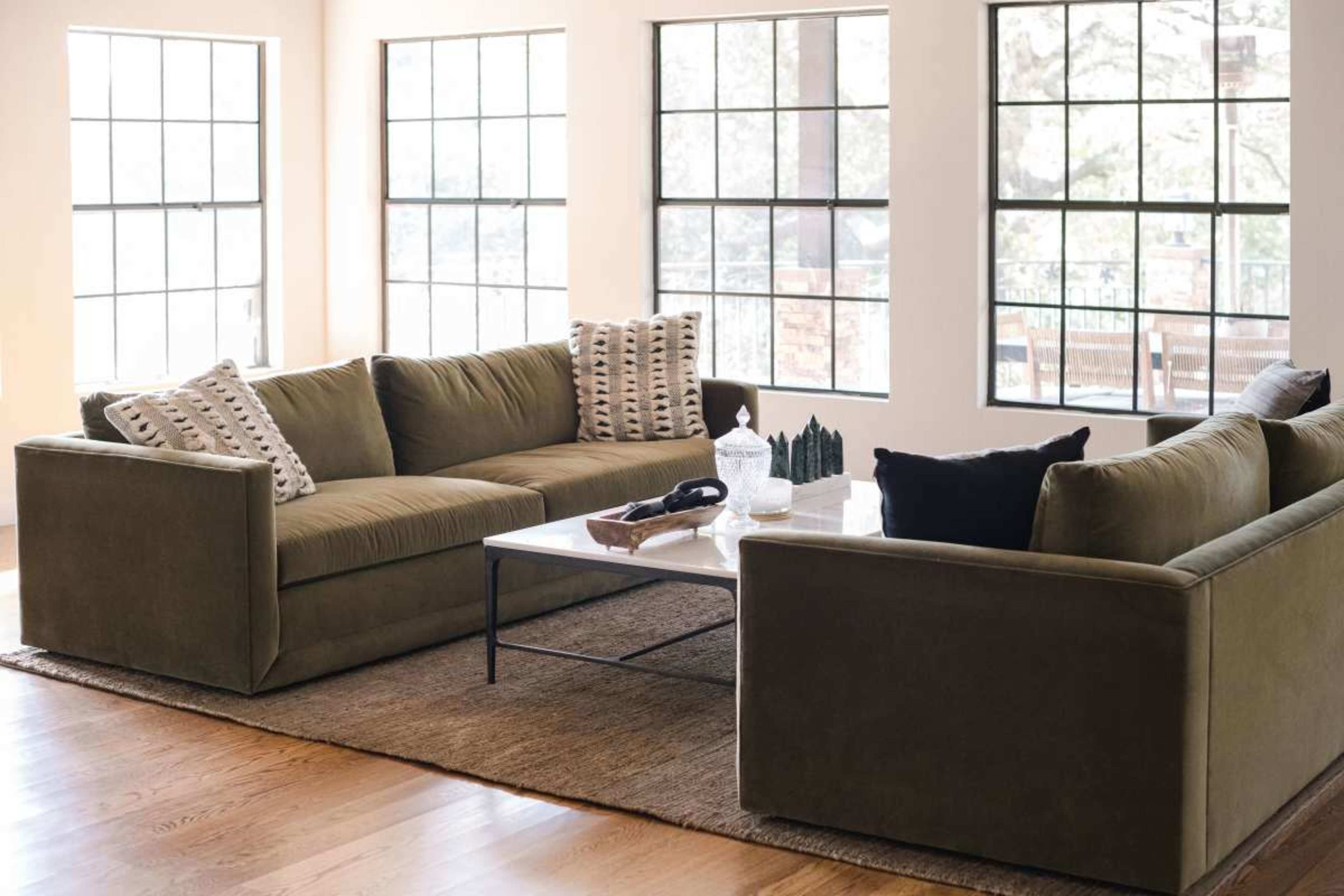 A modern living room features a large, comfortable sectional sofa with decorative pillows, a glass coffee table, and large windows that allow natural light to fill the space.