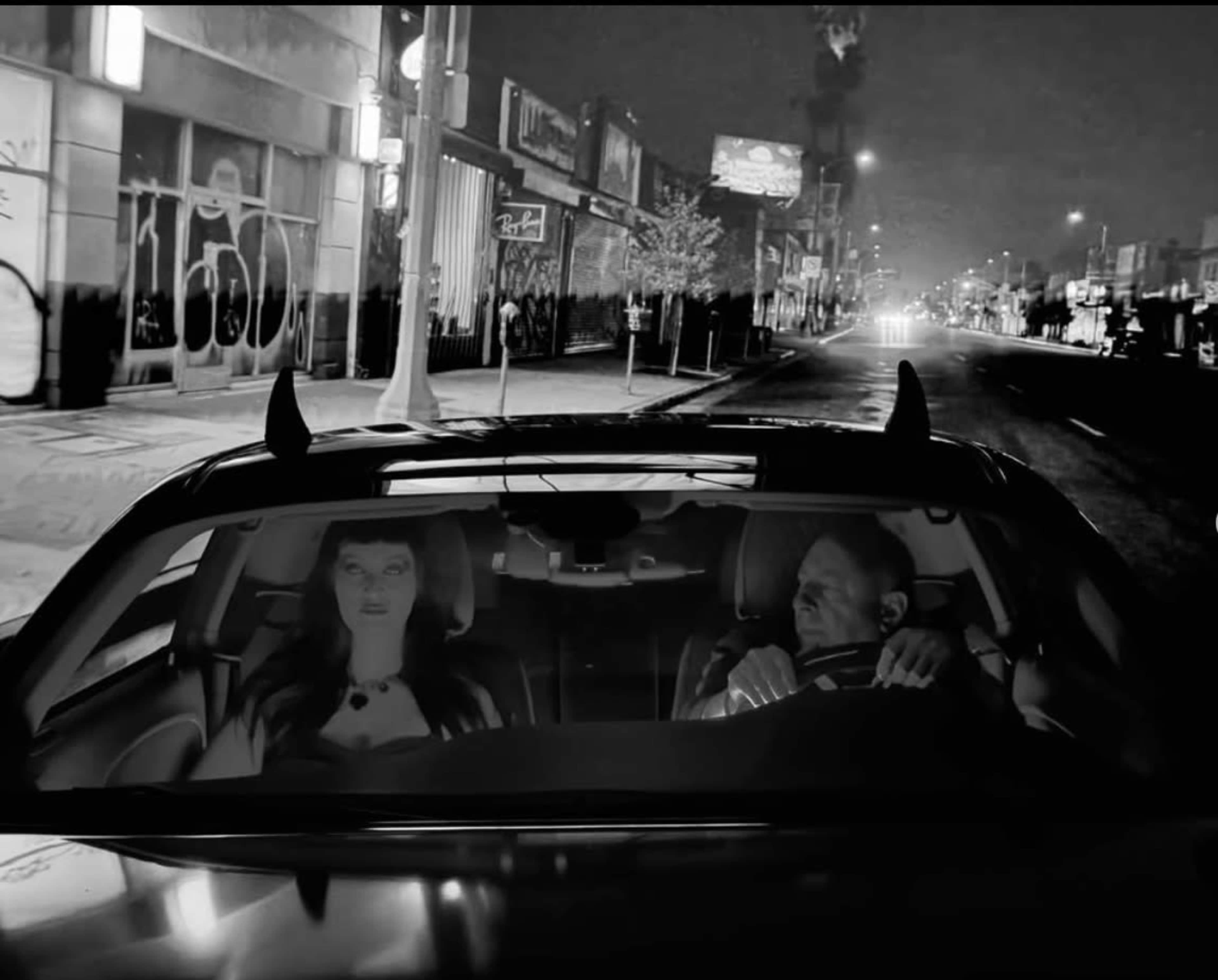 A man and a woman with horns are seated in a car on an empty, dimly lit street at night.