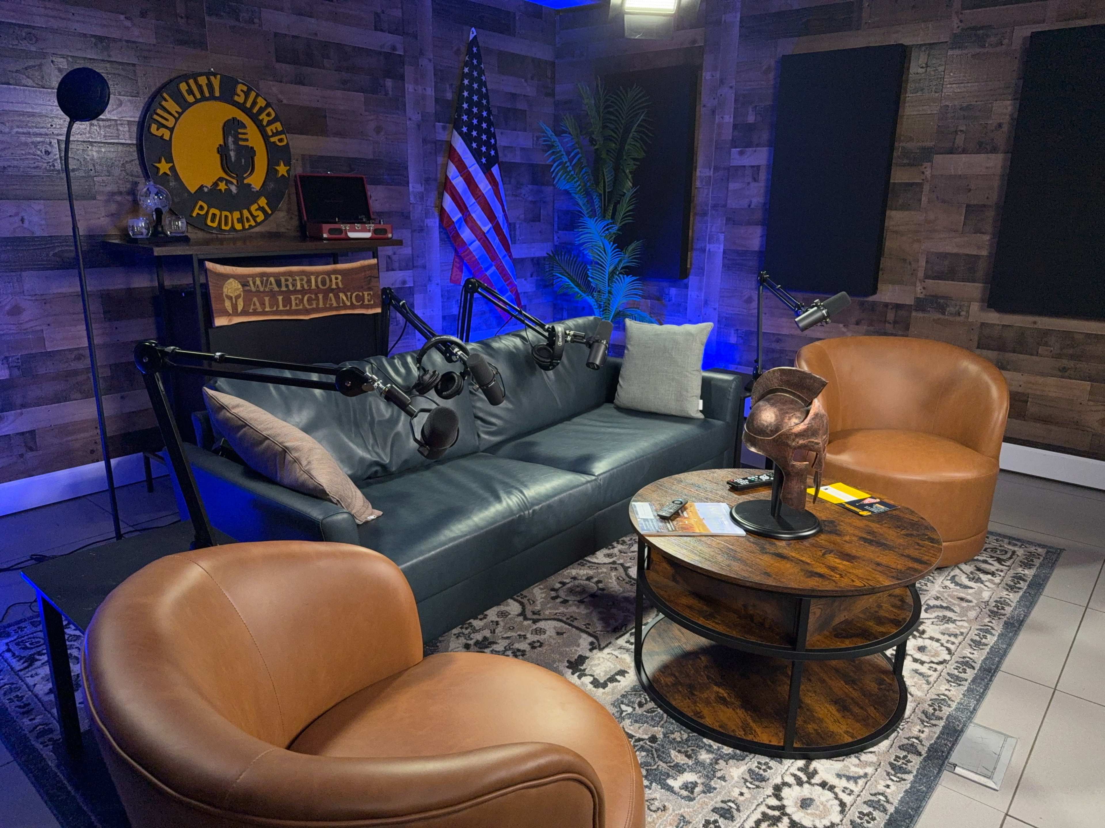 The image shows a podcast studio featuring a dark leather couch, two brown armchairs, a coffee table, a microphone setup, and an American flag in the background.
