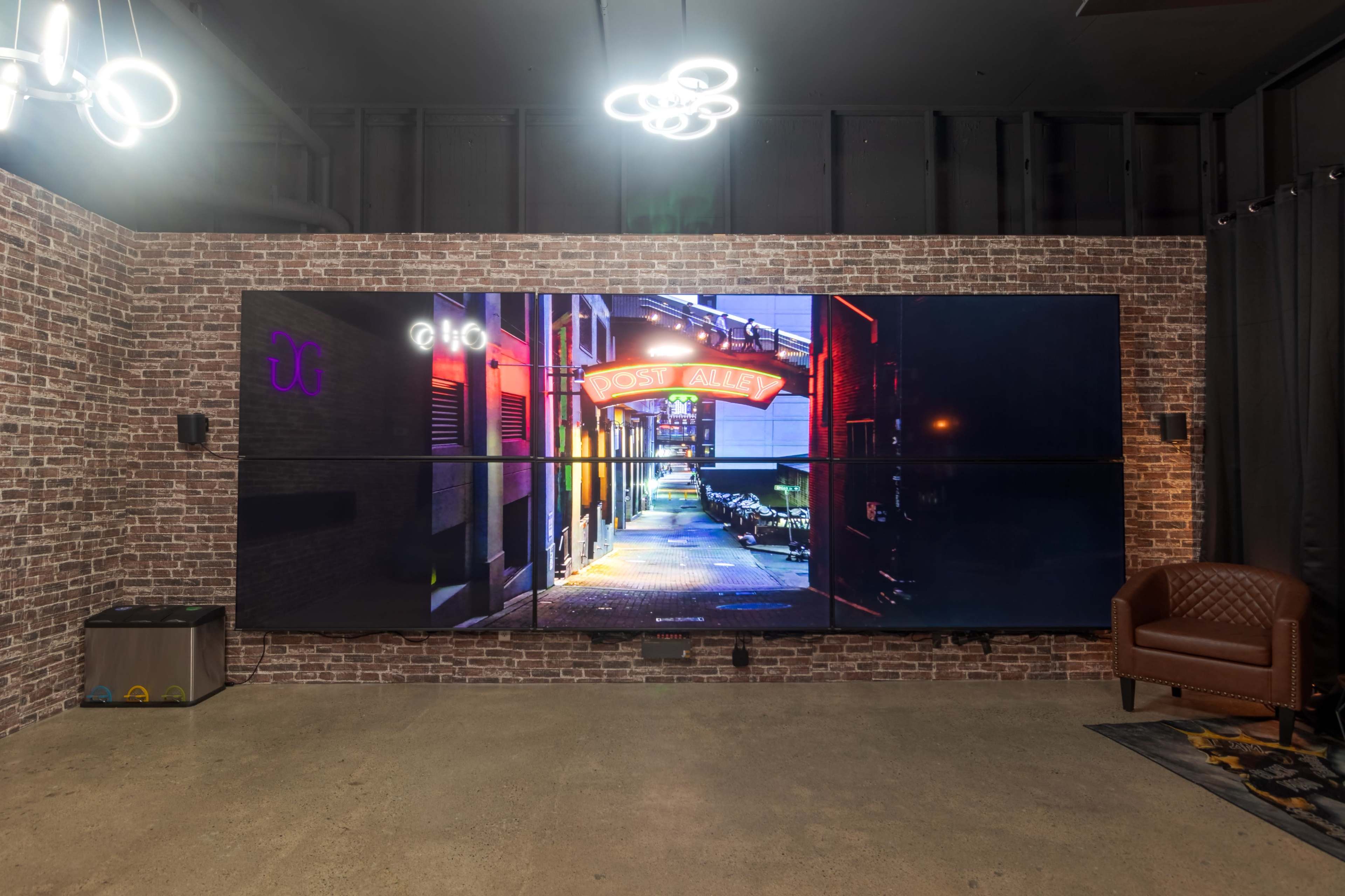The image shows a brick-walled room featuring a large digital display of a brightly lit alleyway, with a visible sign reading "Post Alley."