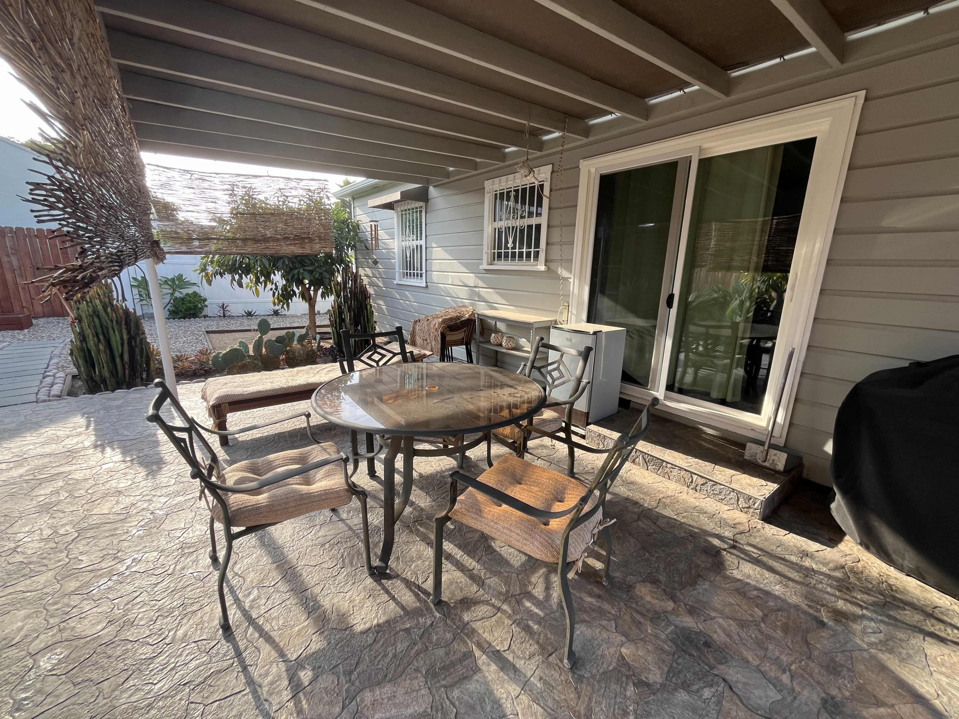 The image shows a patio area with a round table and several chairs, a nearby sliding glass door, and landscaping visible in the background.