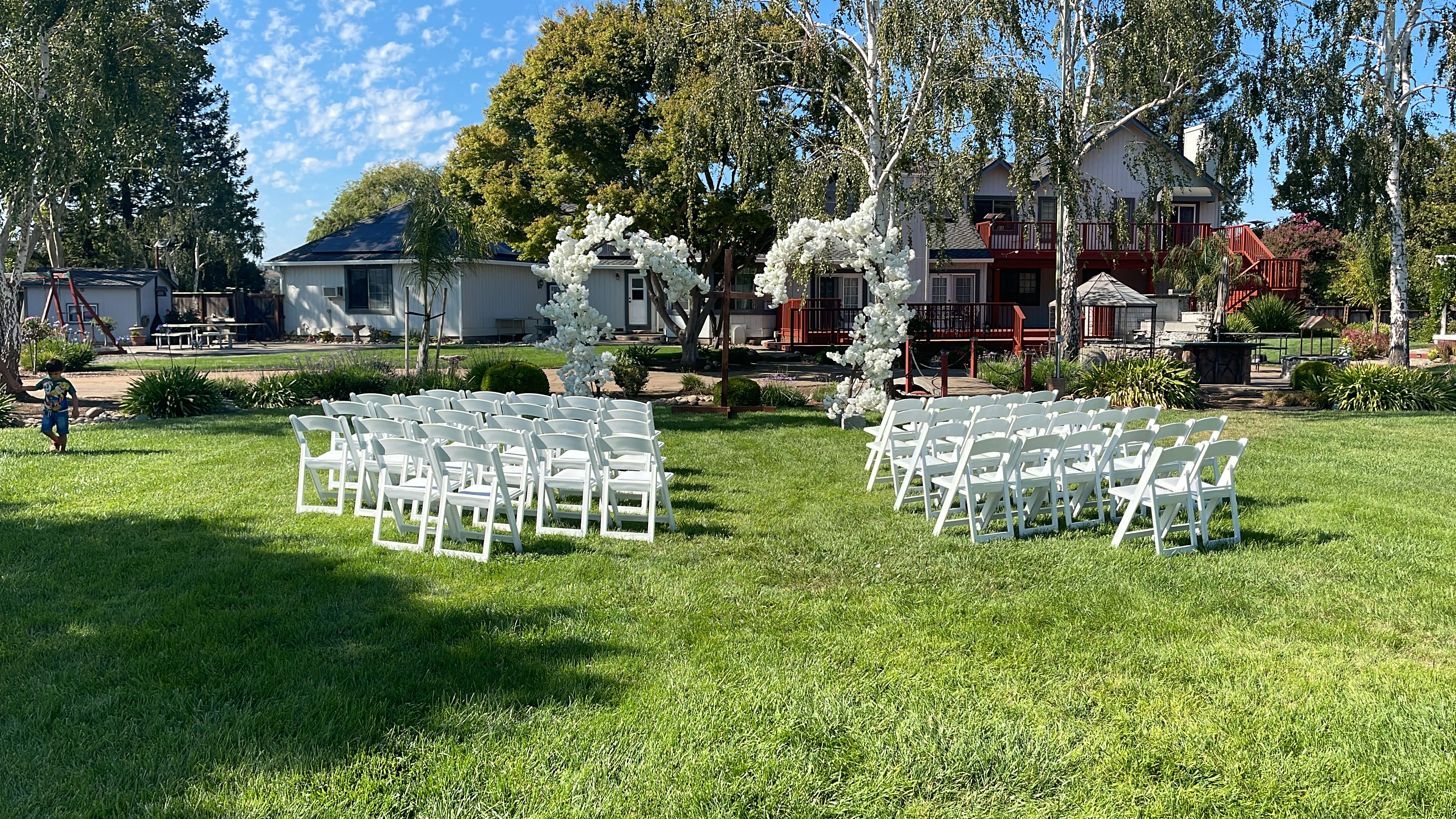 The image shows a neatly arranged outdoor seating area for a ceremony, with white folding chairs set up in two semicircles on a grassy lawn, surrounded by trees and a house in the background.