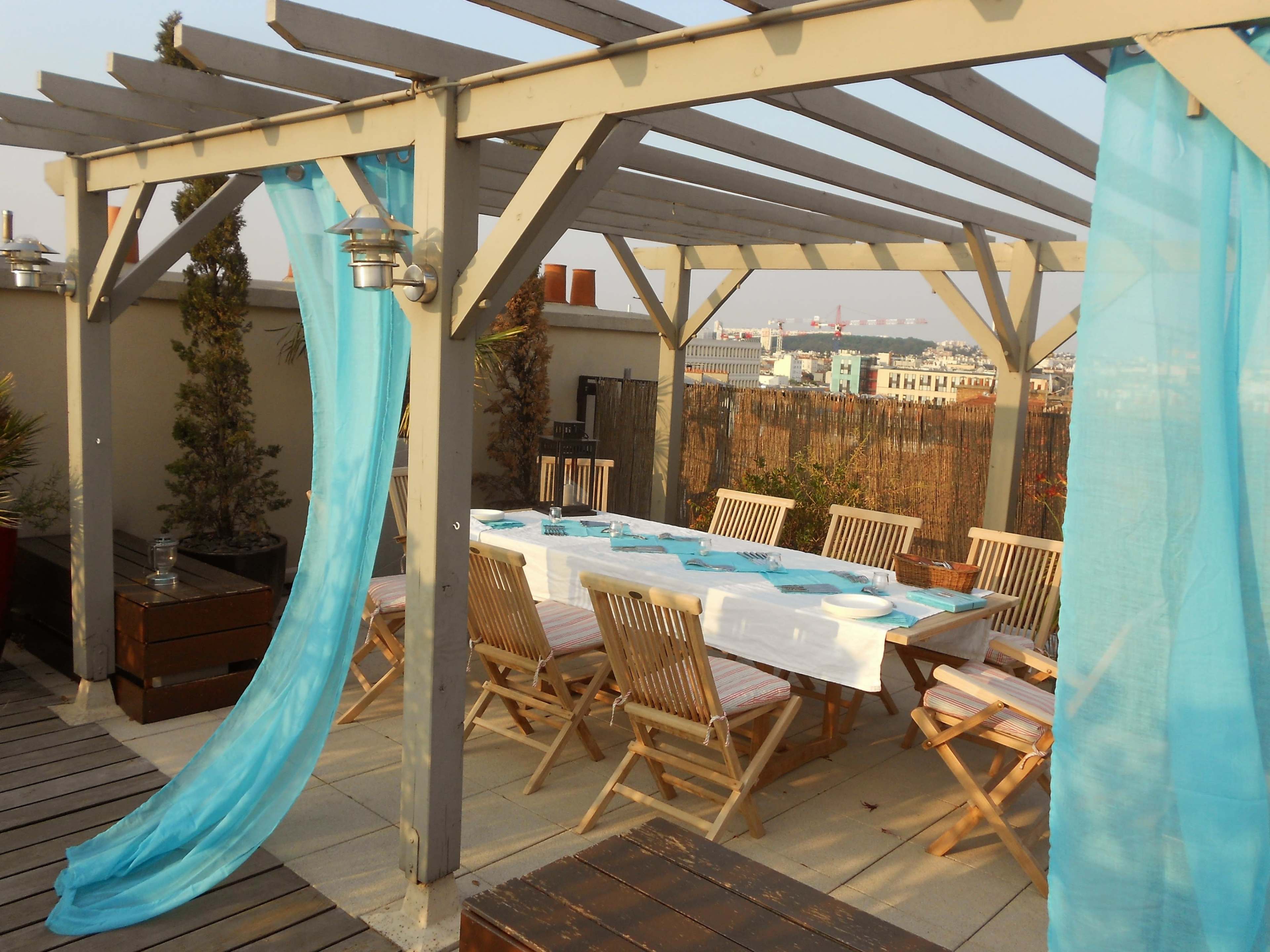 A wooden pergola with turquoise drapes surrounds a dining table set for a meal, accompanied by wooden chairs, on a rooftop terrace.
