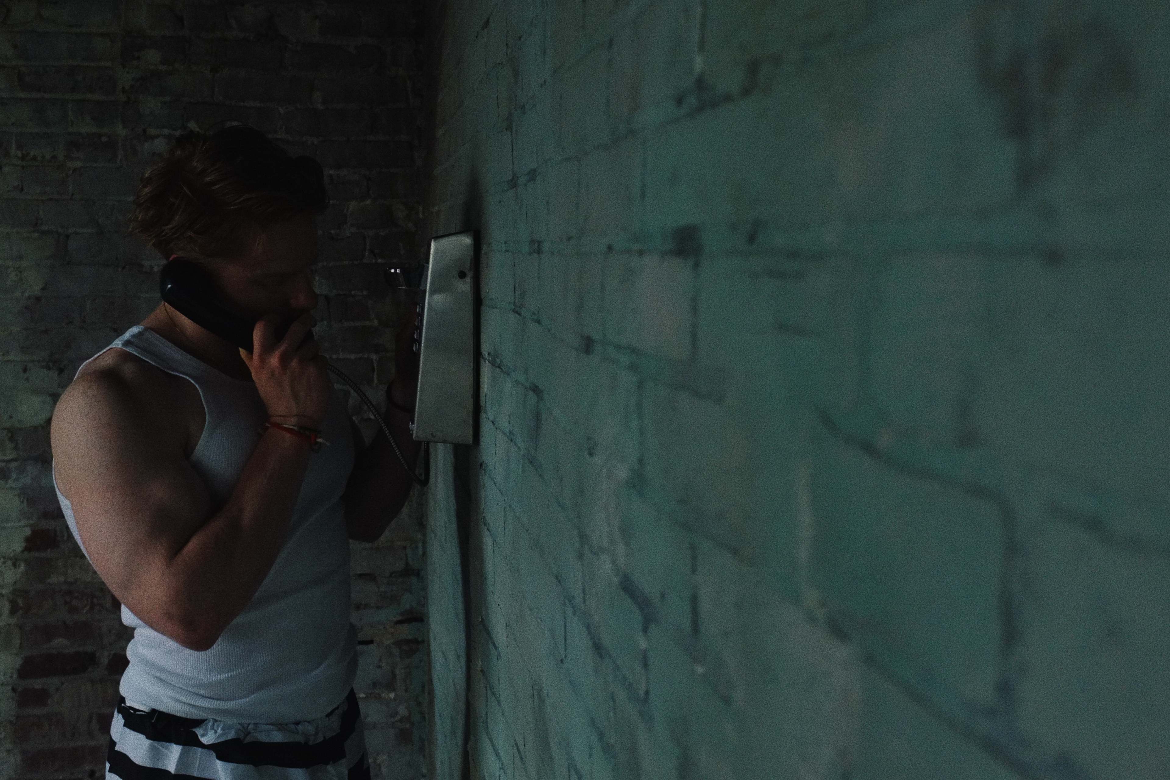 A person is using a payphone against a brick wall in a dimly lit environment.