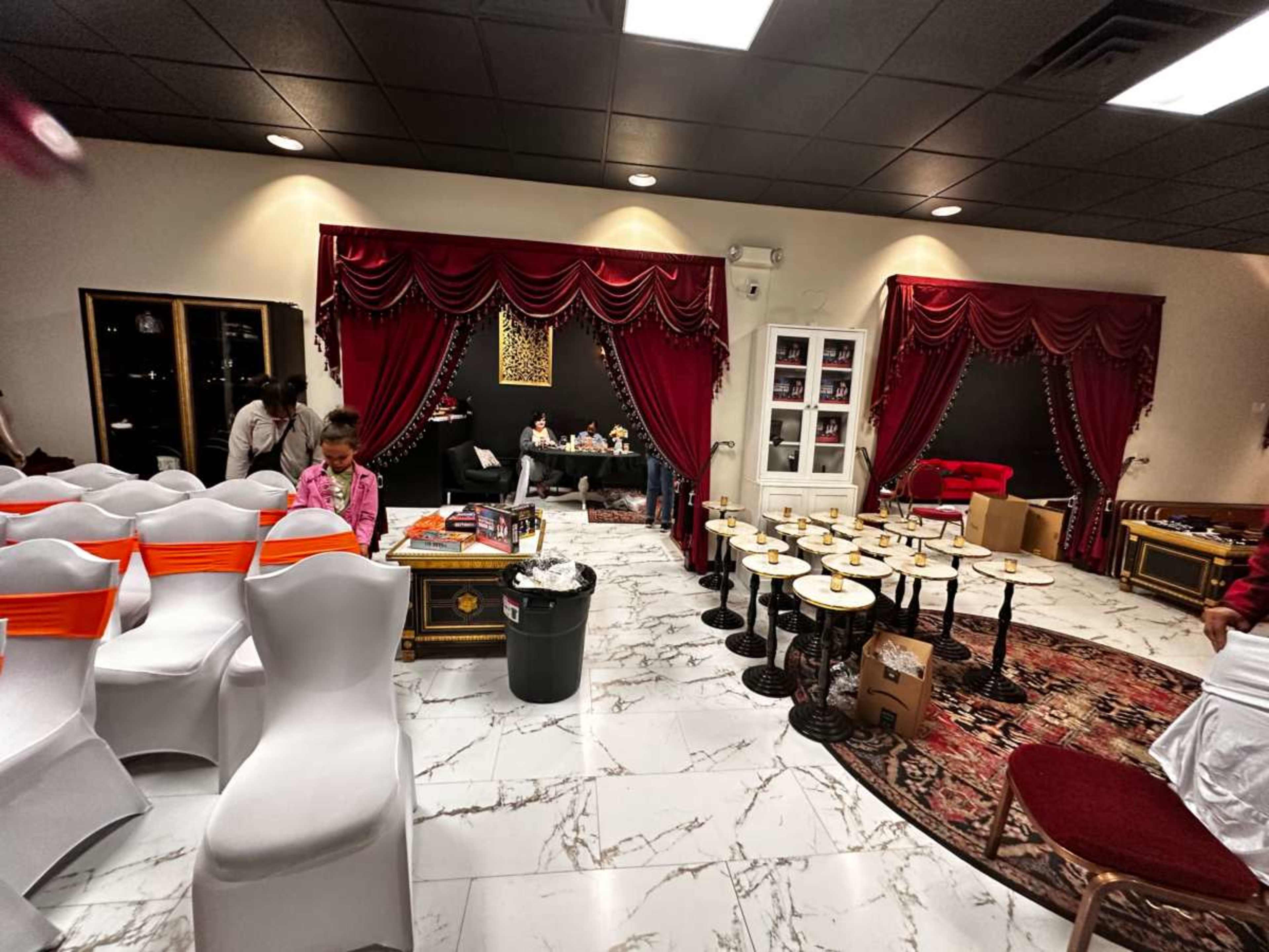 The image shows an event space with white chairs arranged in rows, a red curtained area at the back, and tables set up with boxes and decorations scattered around.