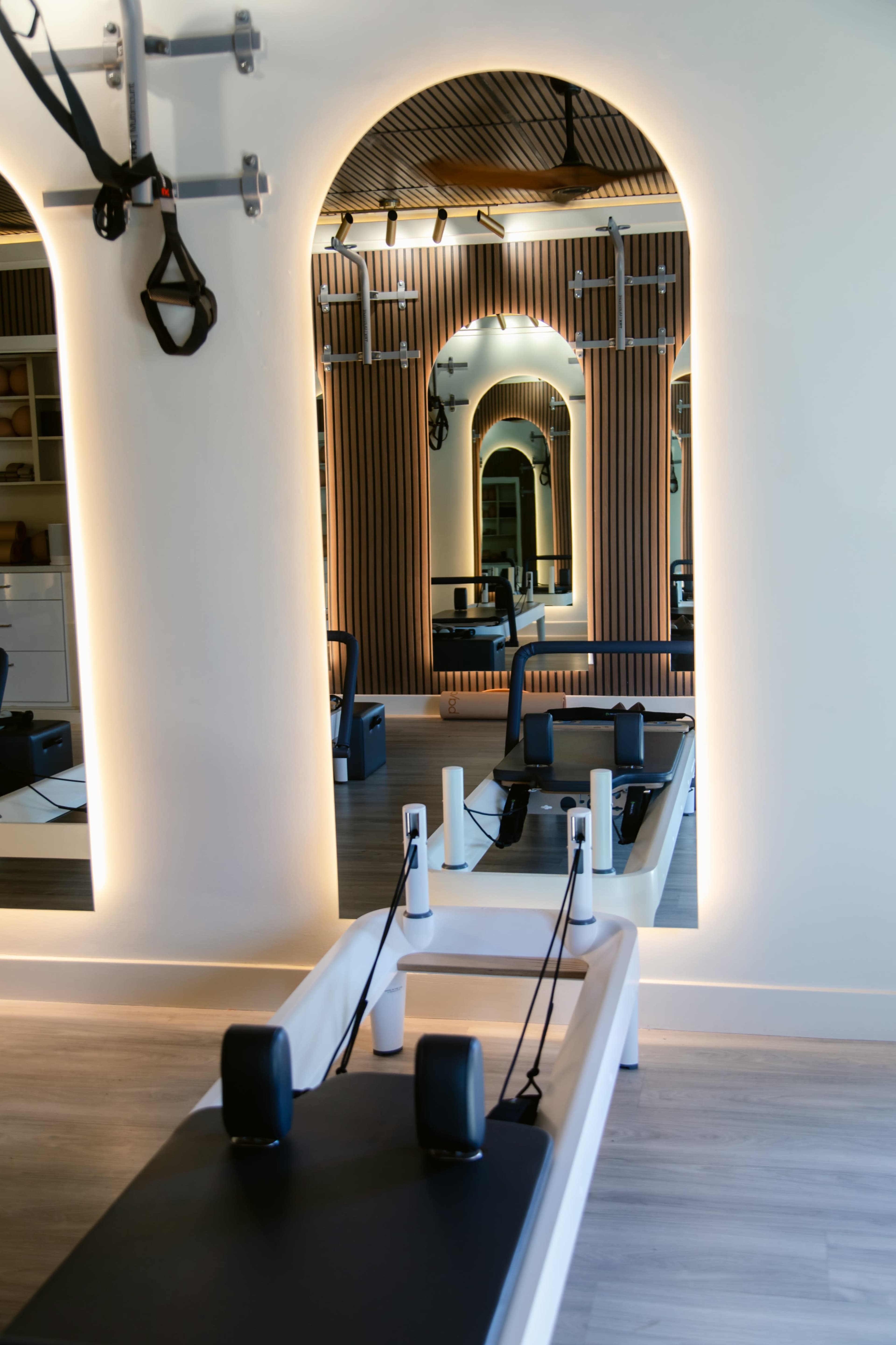 The image shows a modern pilates studio with a reformer machine positioned in front of large mirrors and surrounded by wood paneling.