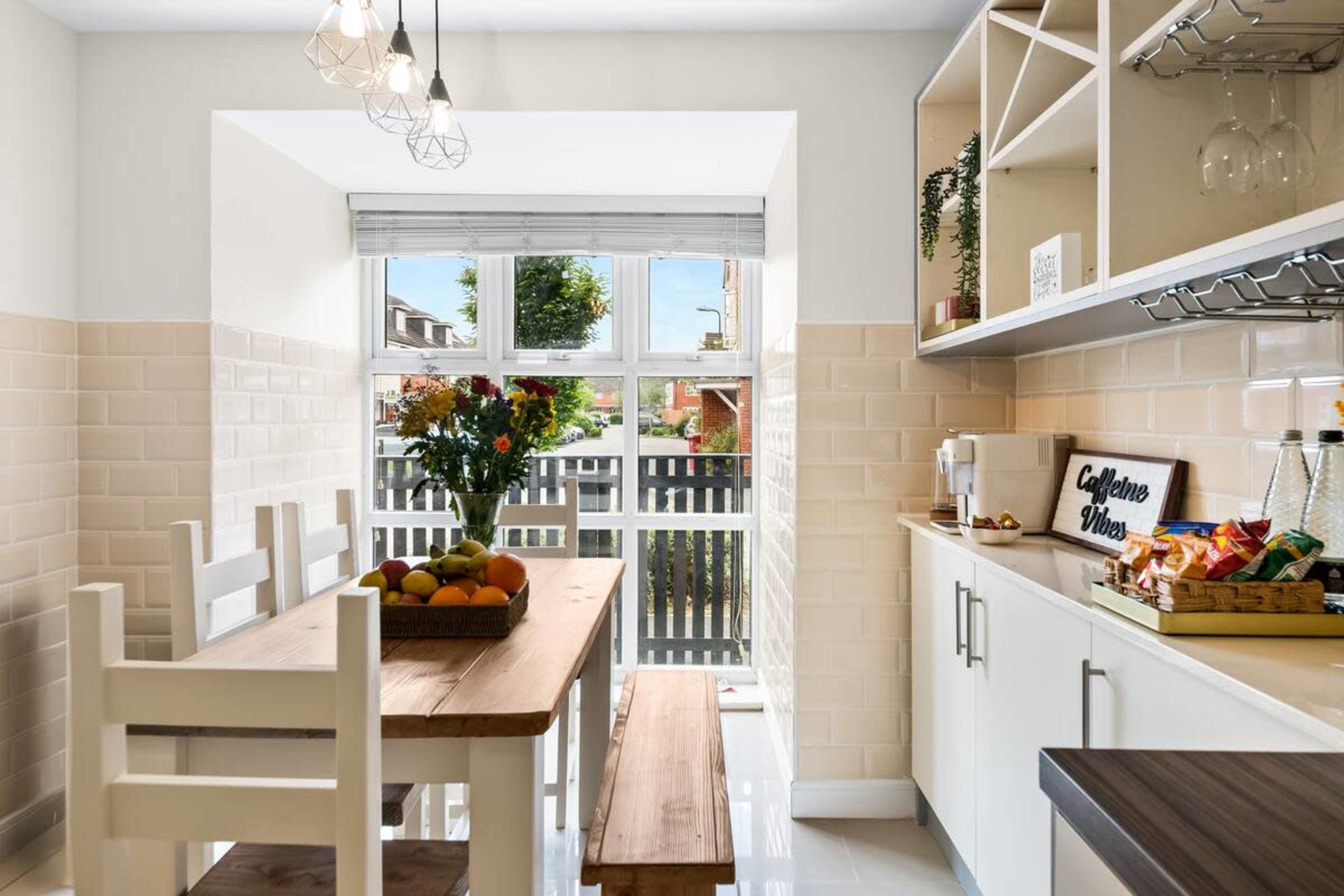 The image shows a modern kitchen area with a wooden table set for dining, surrounded by white chairs and natural light coming through a window.