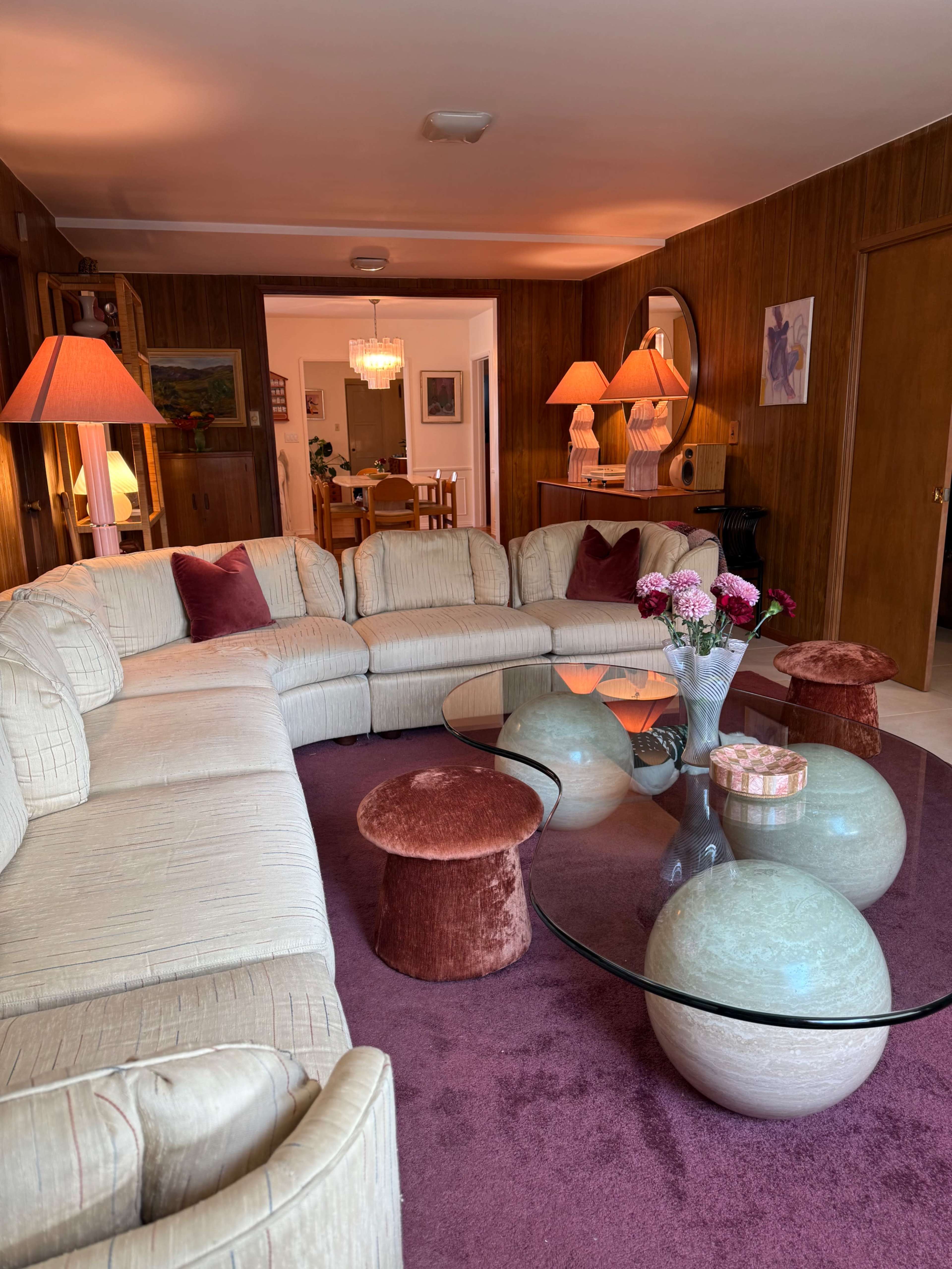 The image shows a living room with a large curved sofa, a glass coffee table, and decorative lights with a warm ambiance.