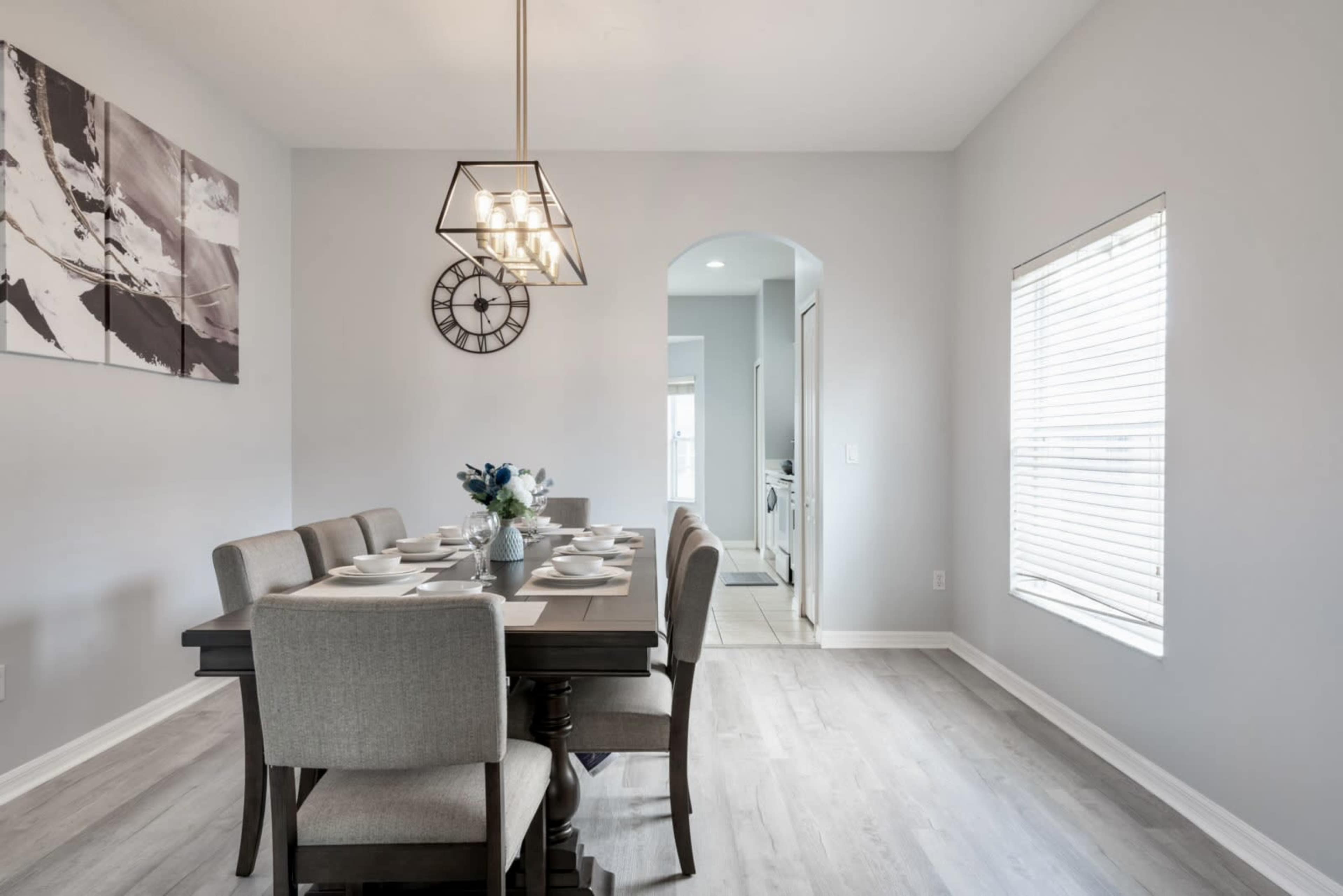 A dining area features a long table set for a meal, with a modern light fixture hanging above, and a clock on the wall beside an archway leading to another room.