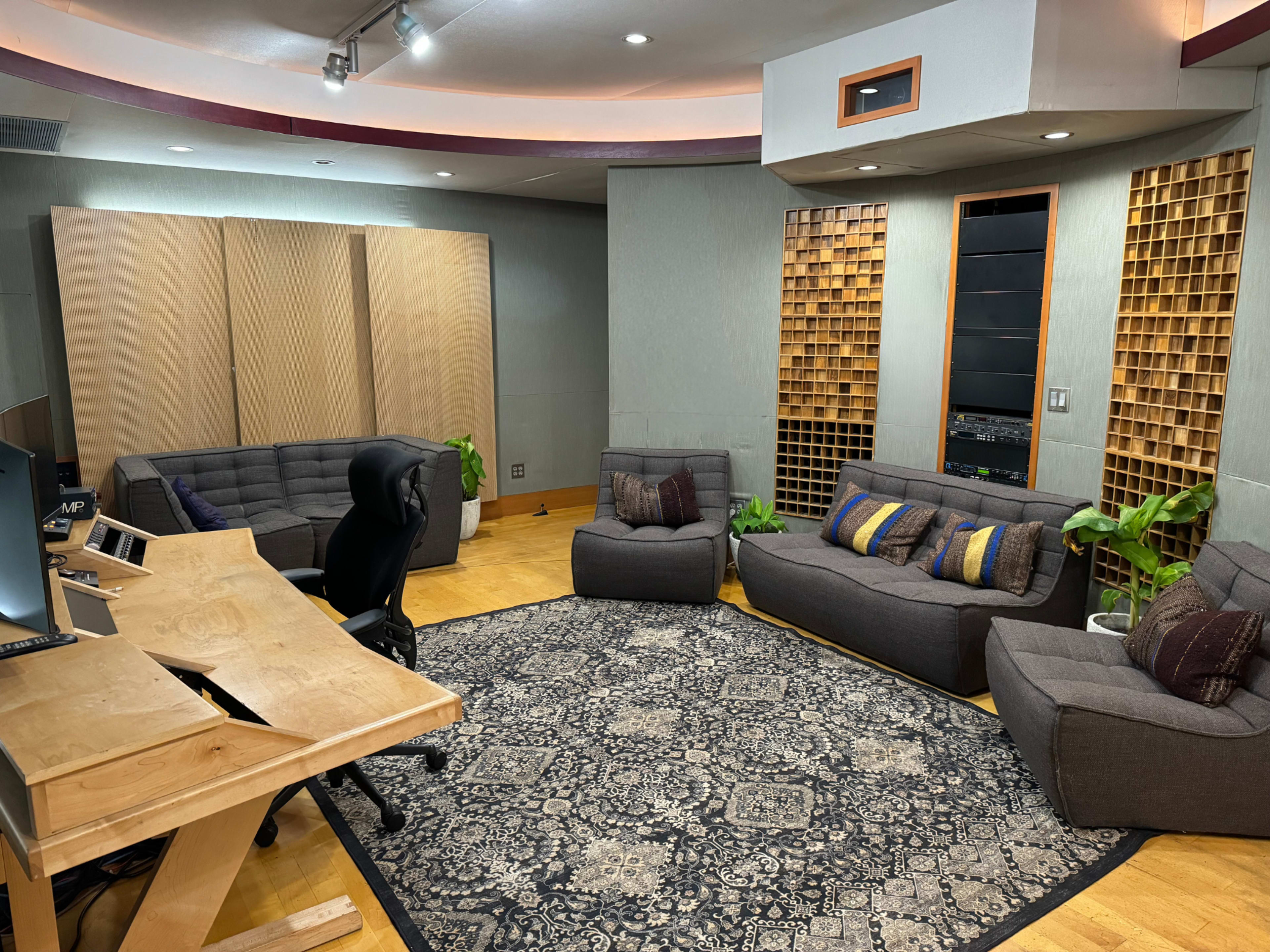 A studio room features a wooden desk, several sofas, and decorative wall panels, with plants placed throughout the space.