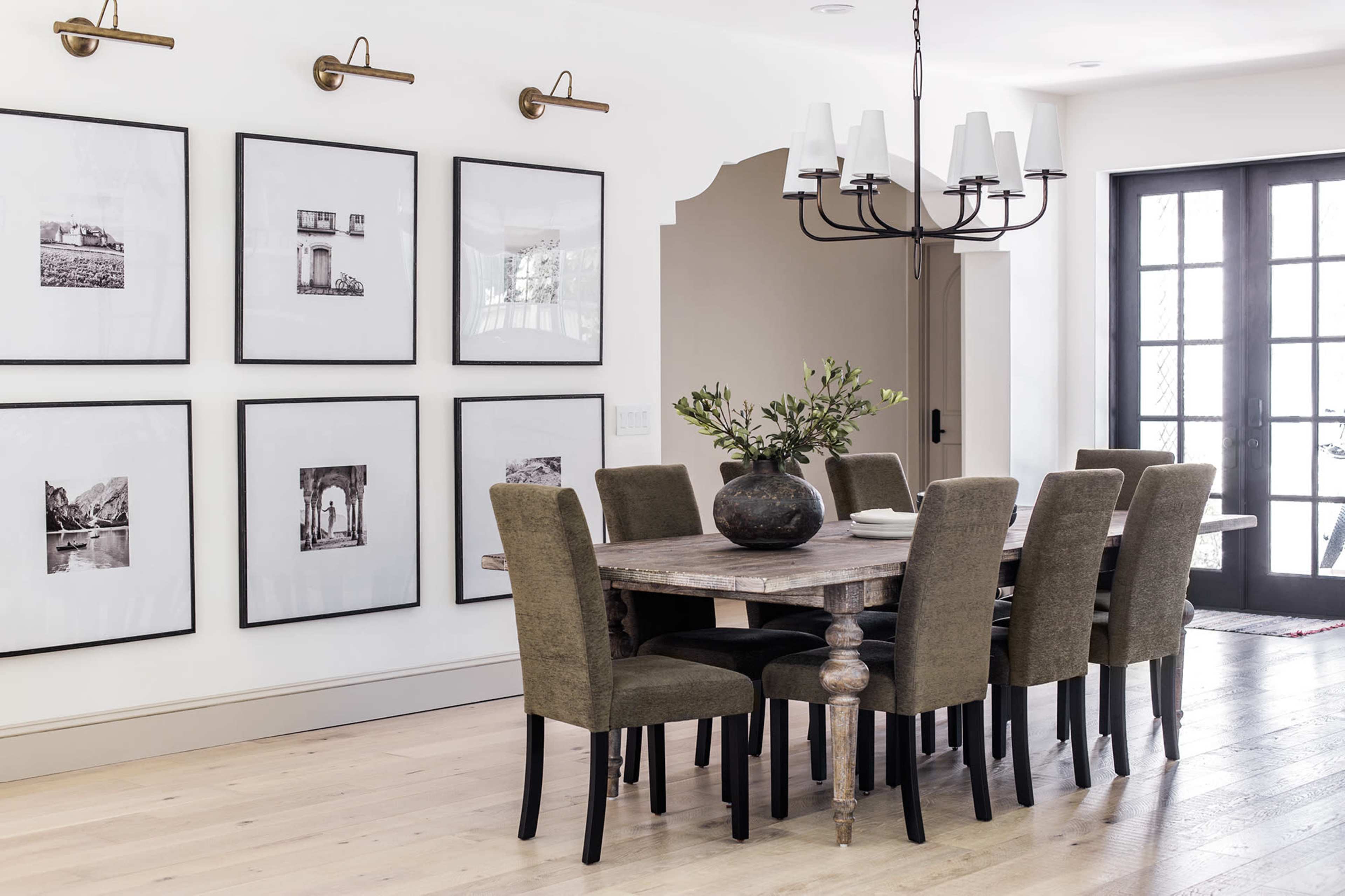 A dining room with a large wooden table surrounded by eight upholstered chairs, complemented by framed black-and-white photographs on the walls and a modern chandelier overhead.