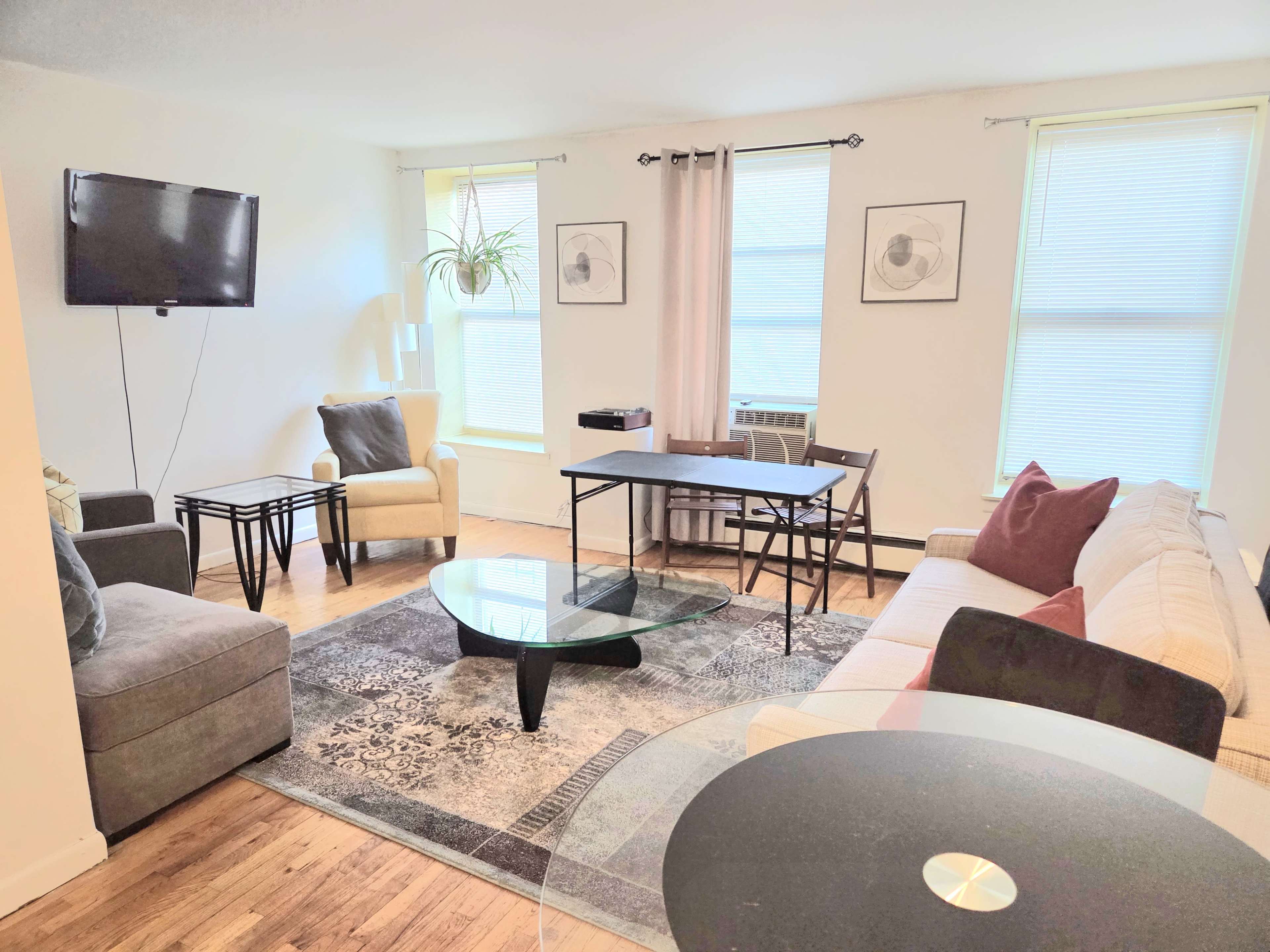The room features a living area with a beige sofa, a glass coffee table, a television on the wall, and two windows, along with a small dining table and chairs.
