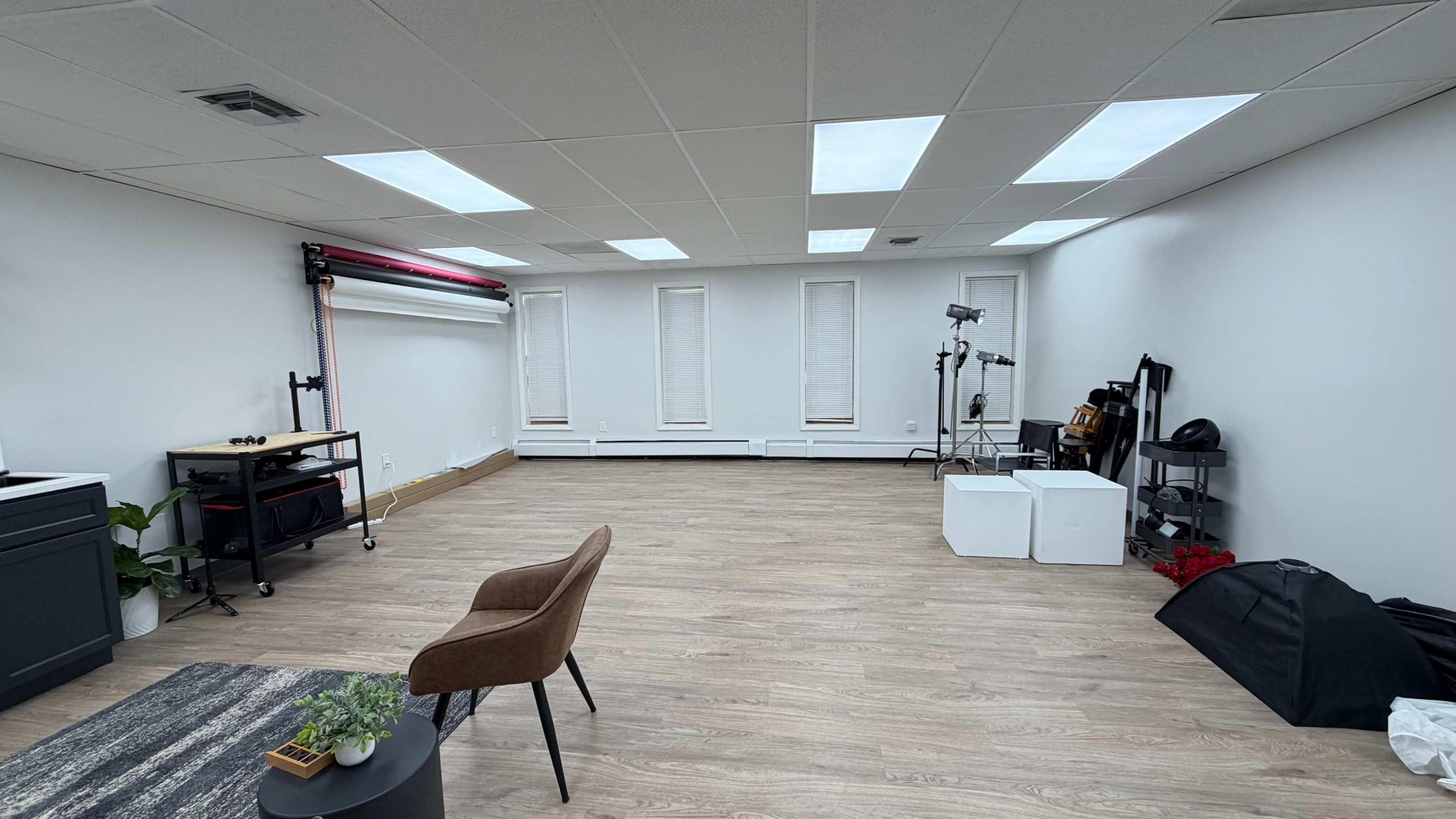 The image shows a spacious, empty room with wooden flooring, several windows, and minimal furnishings including a chair and a couple of small storage units.