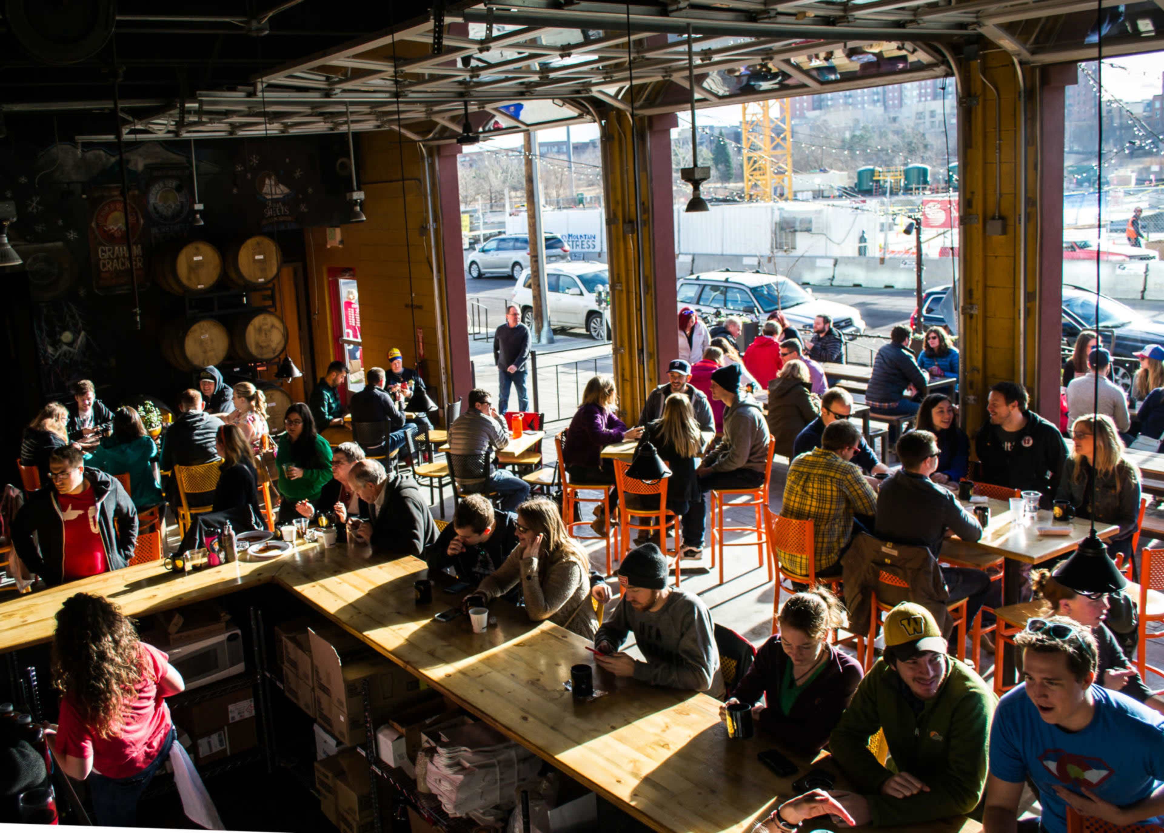 The image shows a bustling café with patrons seated at wooden tables, enjoying their drinks and conversations while large windows display a view of the street outside.