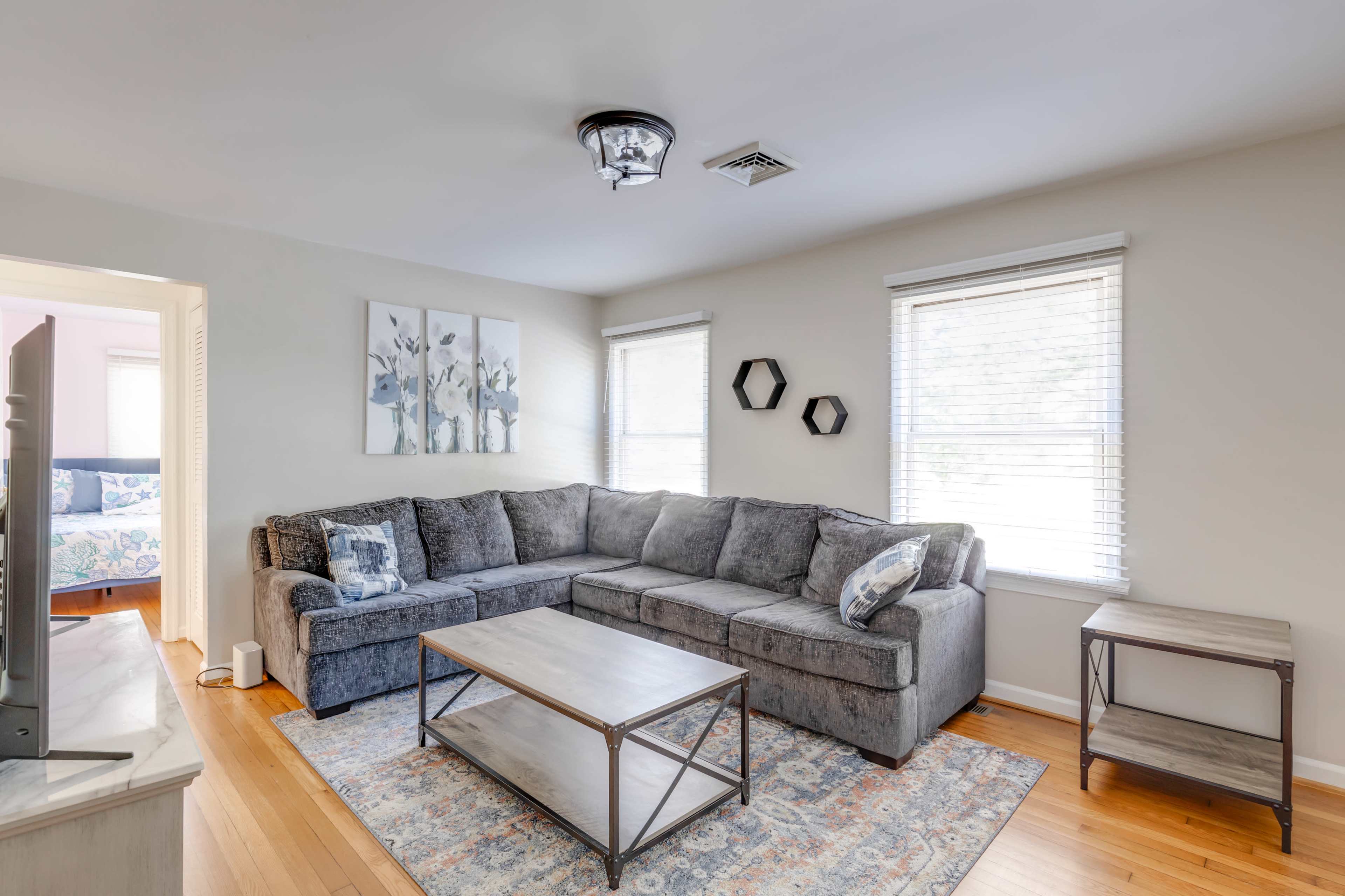 A living room featuring a gray sectional sofa, a rectangular coffee table, and two windows with white blinds.