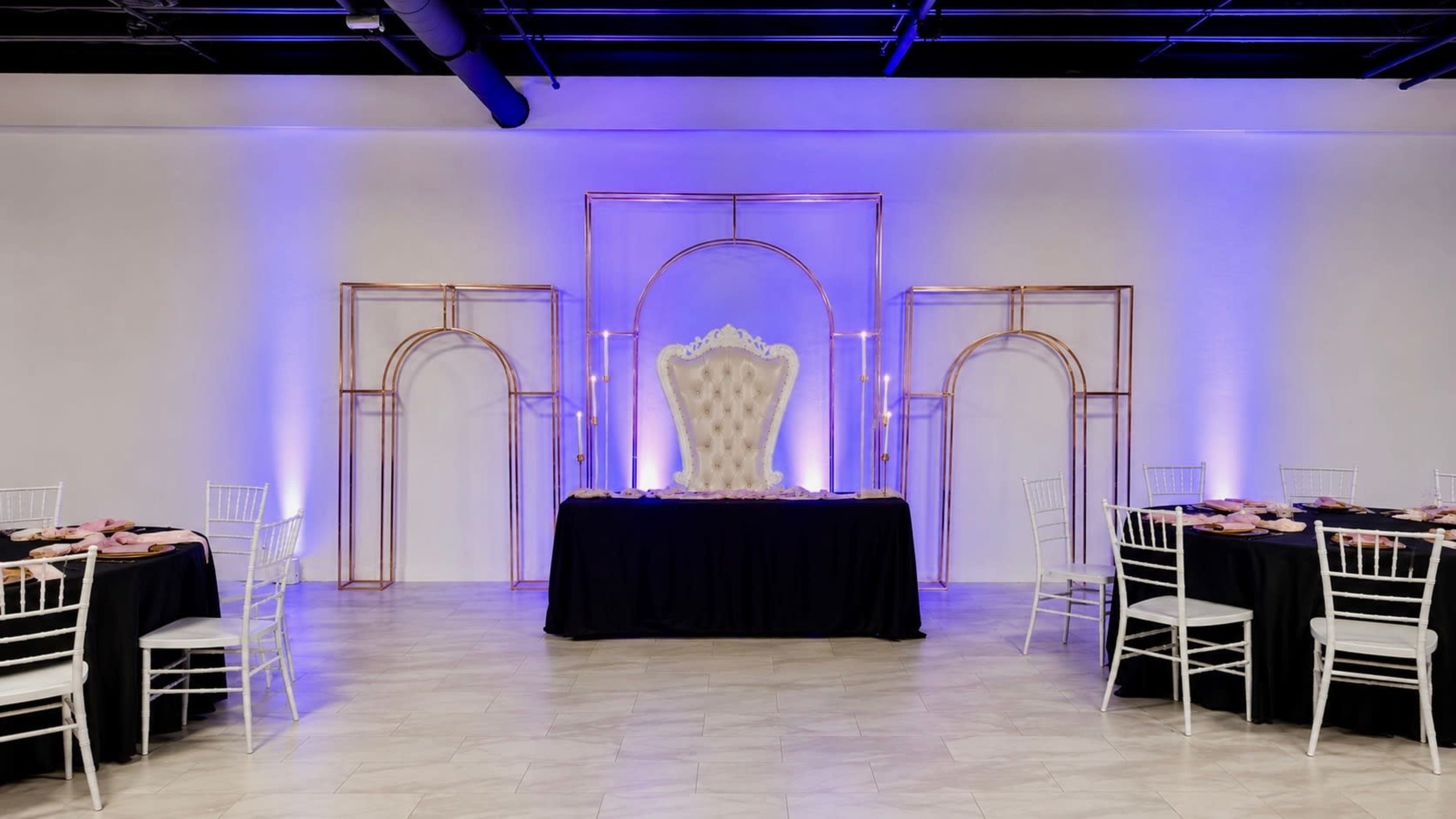 A lavish event space features a grand white throne center stage, flanked by gold arch decorations and illuminated with purple lighting, surrounded by elegantly set tables.