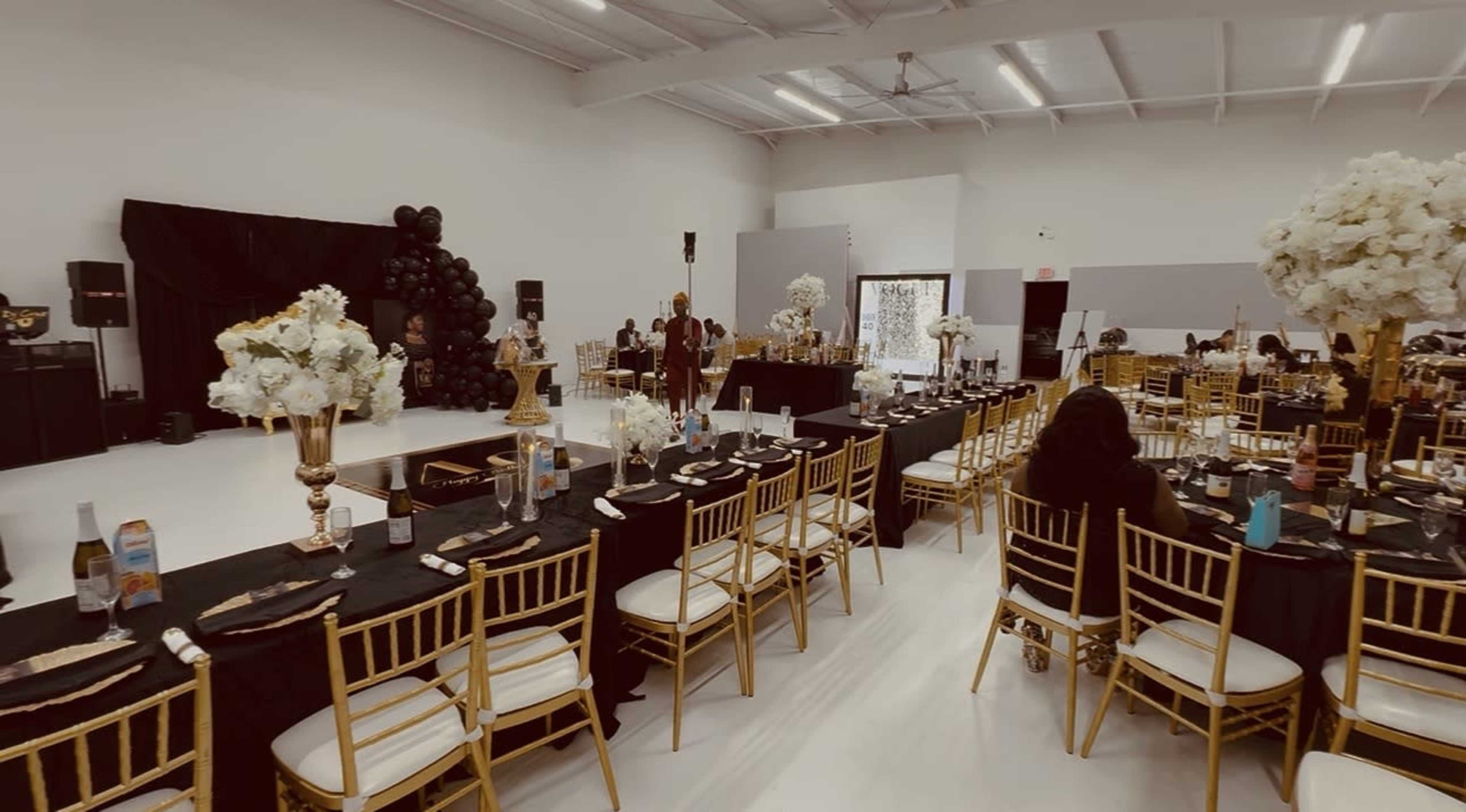 The image shows a spacious event hall set up for a formal gathering, featuring black tables adorned with gold chairs and floral centerpieces.