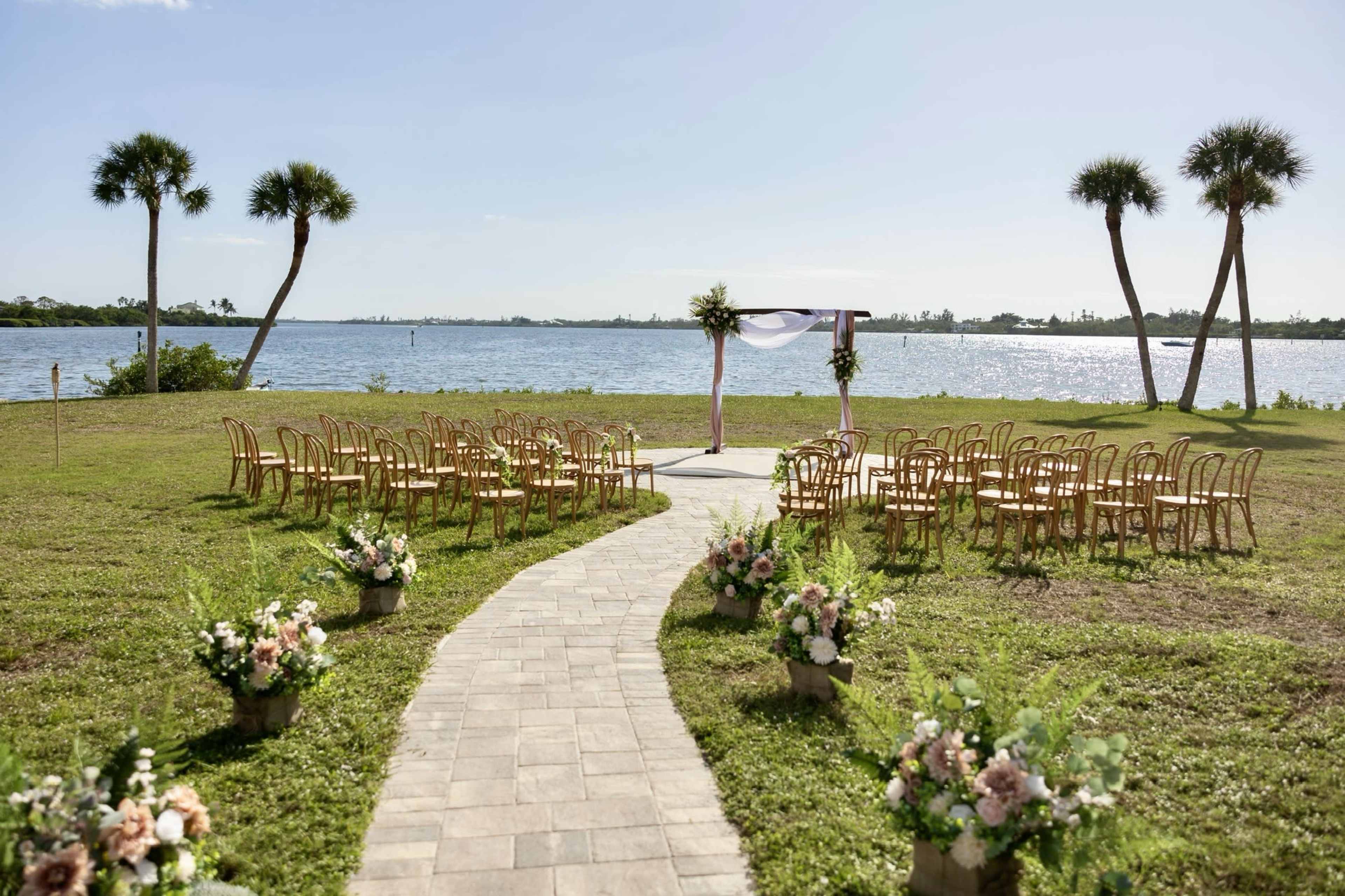 The image shows a wedding ceremony setup by a waterfront, featuring a decorated arch, rows of chairs, and floral arrangements along a paved path.
