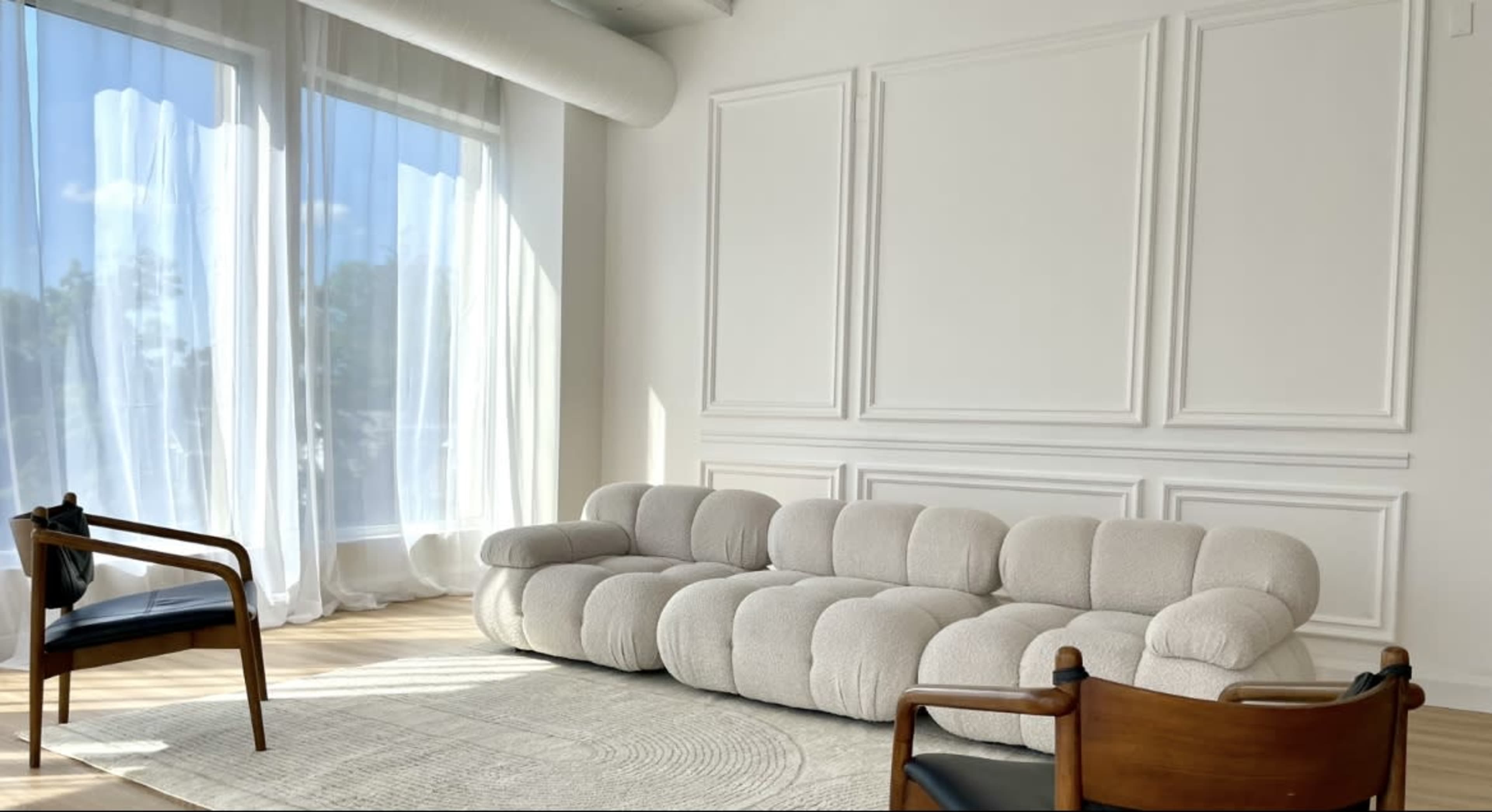 A modern living room with a large, curved beige sofa, two wooden chairs, and large windows covered with sheer white curtains.