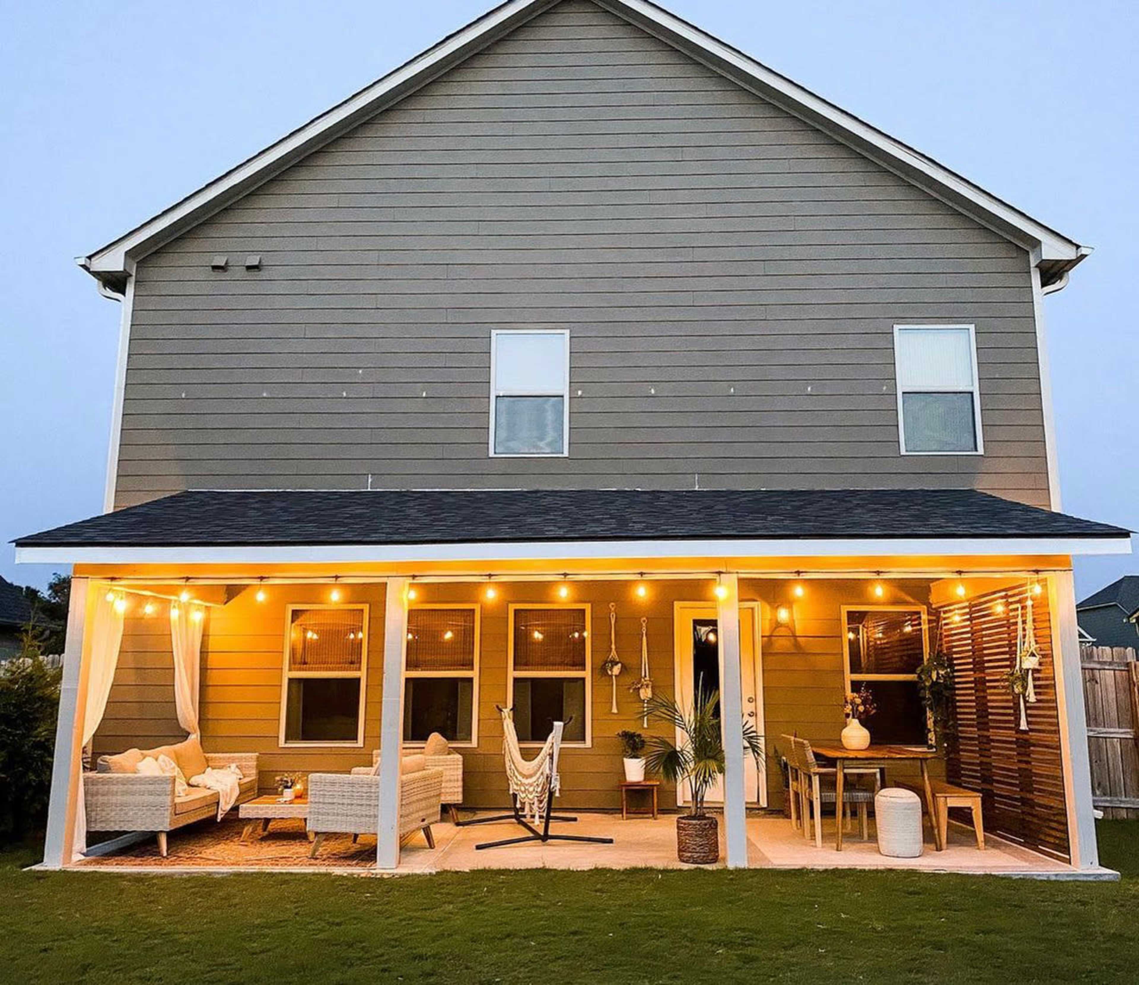 The image shows a lighted porch area attached to a two-story house, featuring seating arrangements, a hammock, and potted plants.