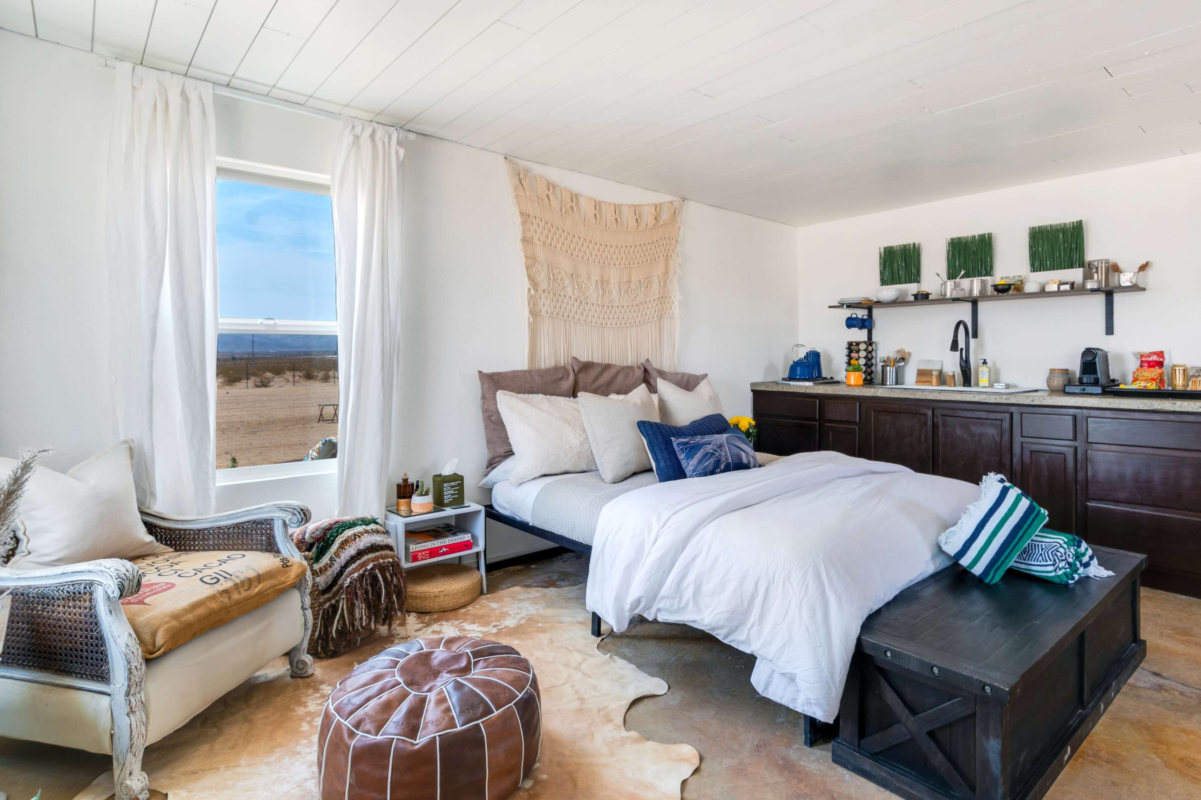A minimalistic bedroom featuring a bed with white linens, a macramé wall hanging, and a small kitchenette with a wooden counter against a window that offers a view of the desert landscape.